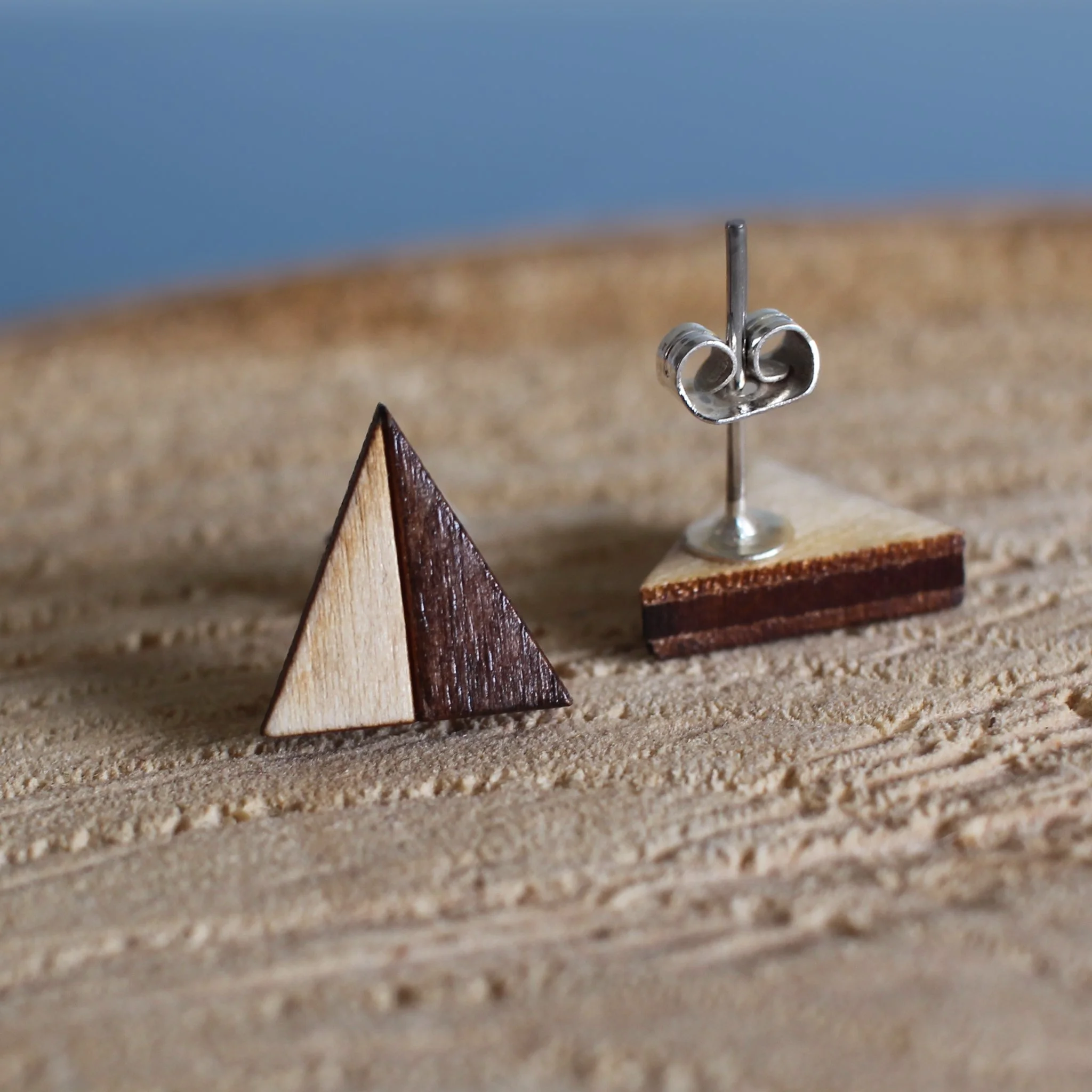 Wooden stud earrings displayed on a log slice. One stud faces the camera, showing a triangle design. The other lies face down, displaying a resin set silver post and butterfly scroll back.