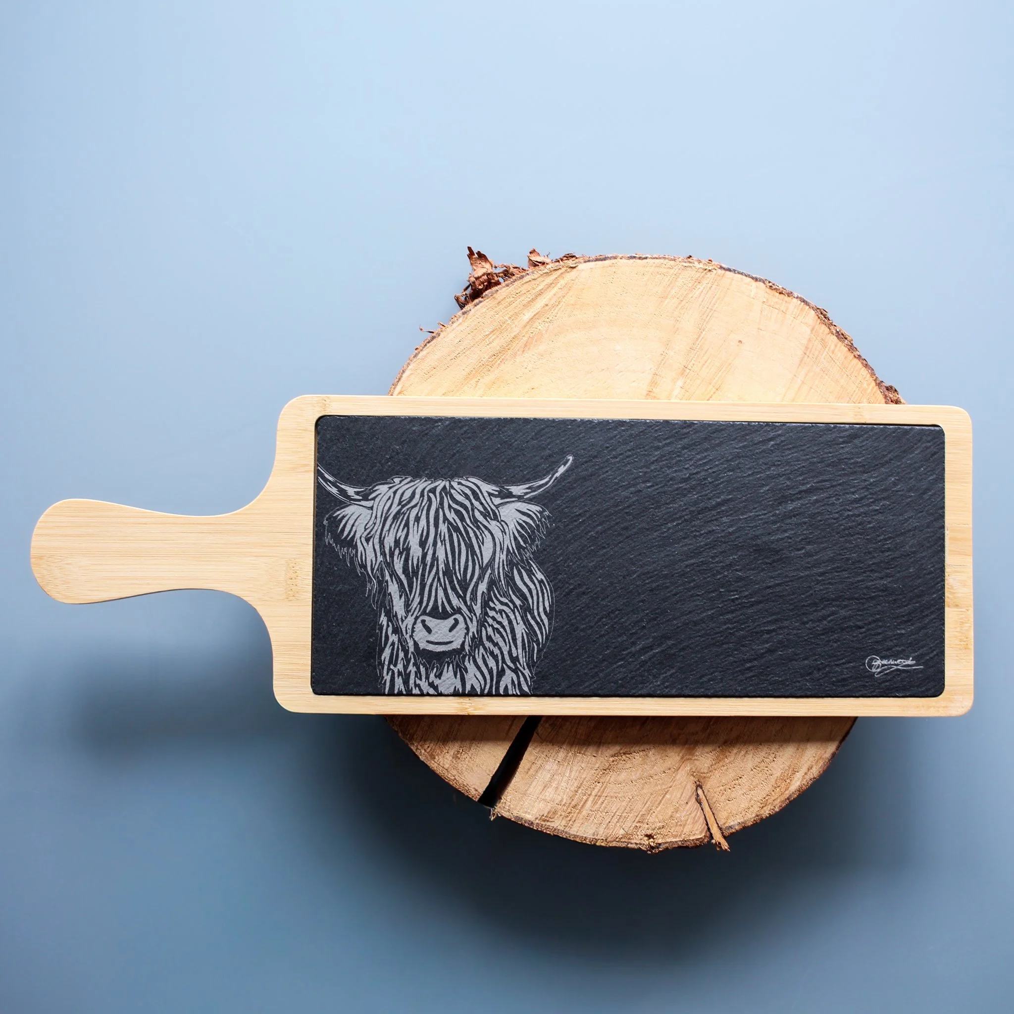 Hand drawn highland cow illustration, engraved onto slate insert, set inside bamboo tray platter. Displayed on log slice on blue background.