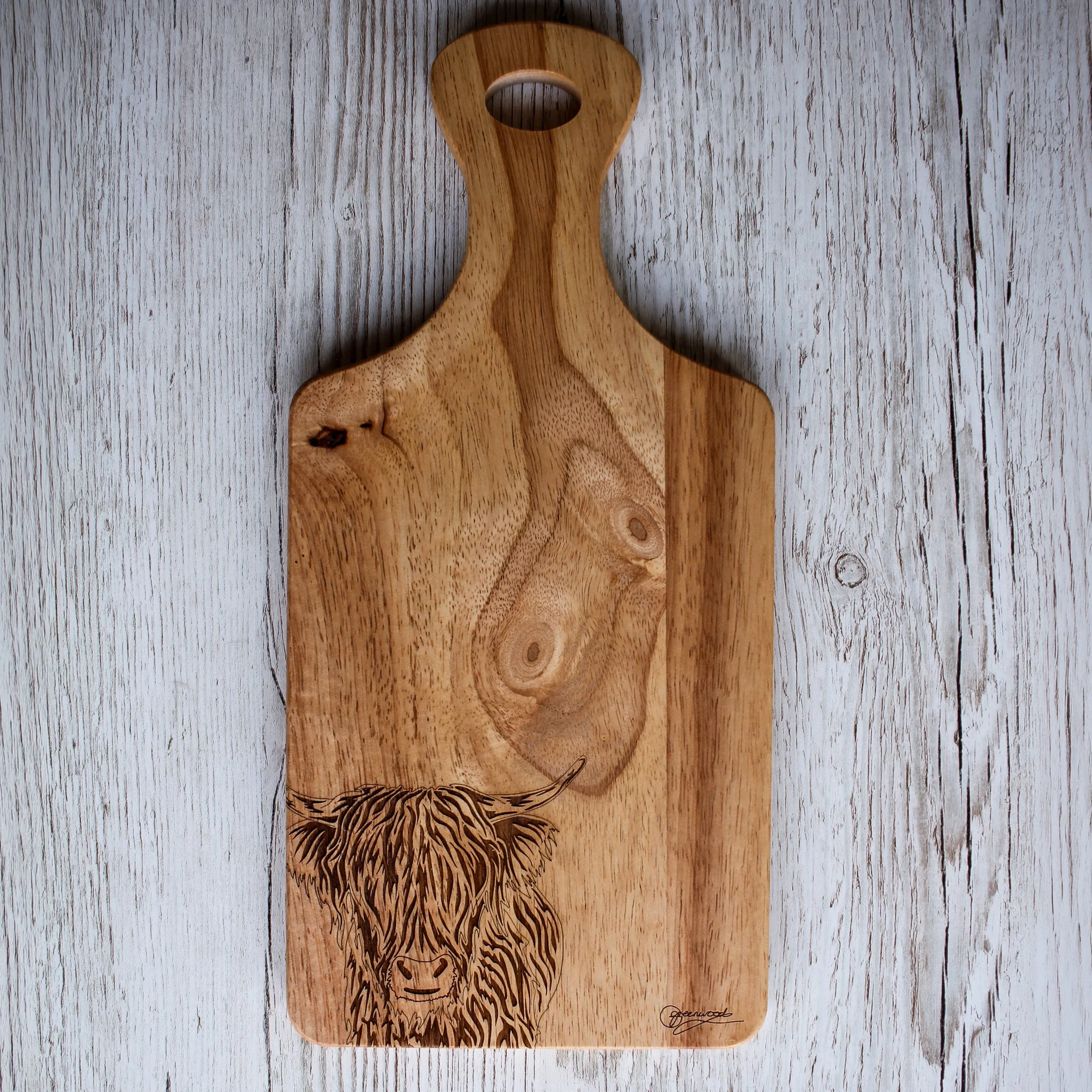 Wooden paddle chopping board with hole cut in tapered end and highland cow art engraving. Displayed on whitewash wood background.