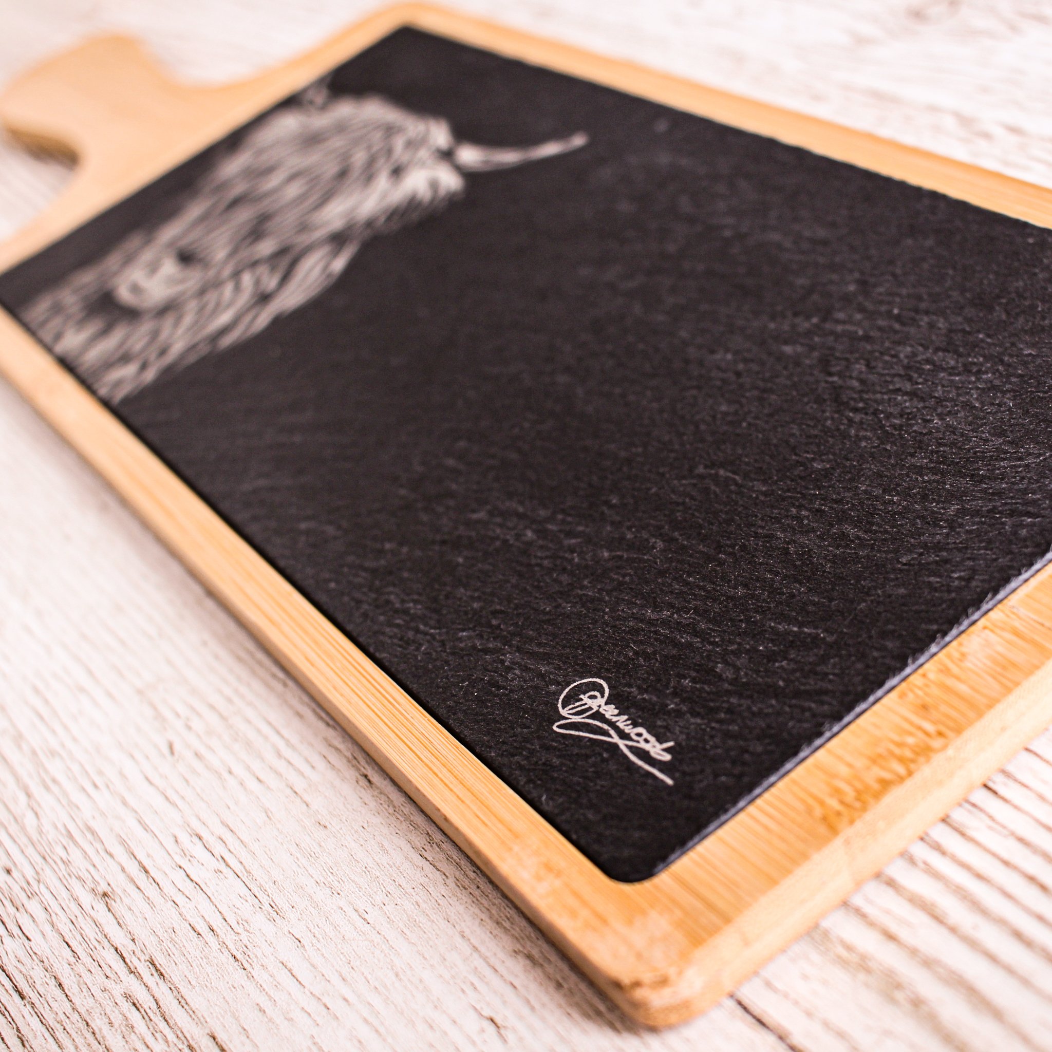 Slanted shot showing highland cow serving platter, focus on the artist signature whose artwork is engraved onto the slate in this kitchenware piece.