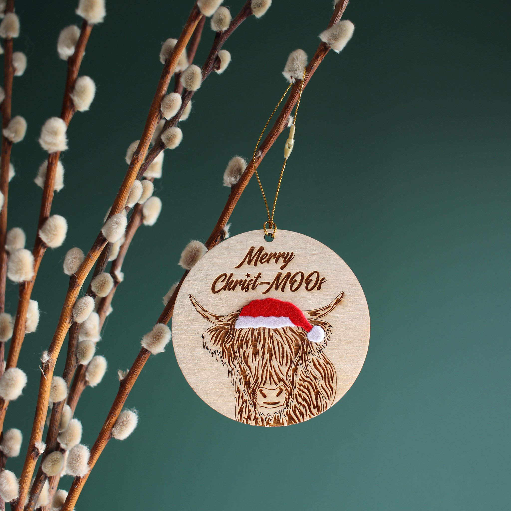 Highland cow Christmas tree ornament hanging from pussy willow branch. The ornament is laser cut and engraved from wood and hand finished with velvet accents. It reads “Merry Christ-MOOs”.