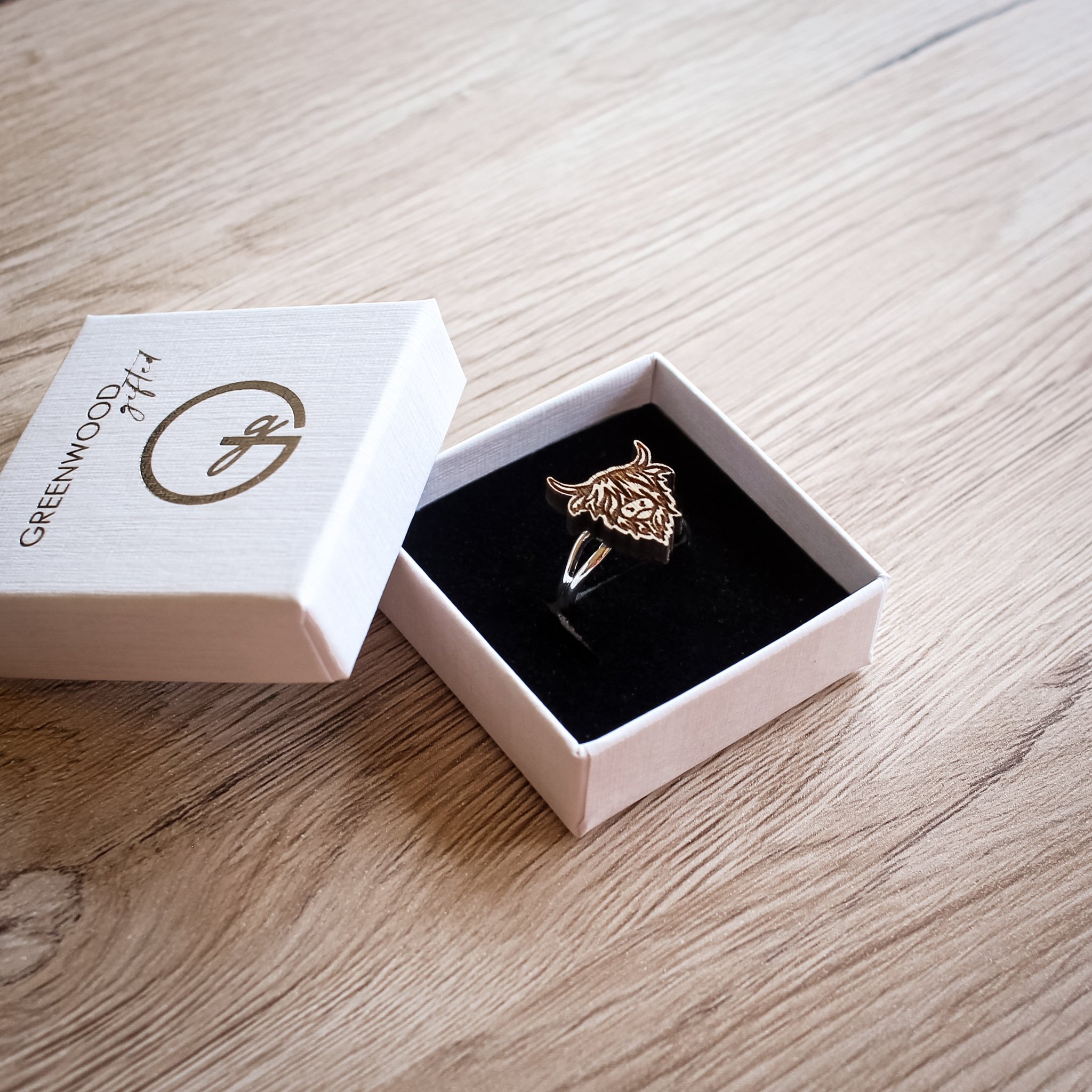 A wooden Highland Cow ring with engraved detail displayed in a white Greenwood Gifted jewellery box on a wooden surface