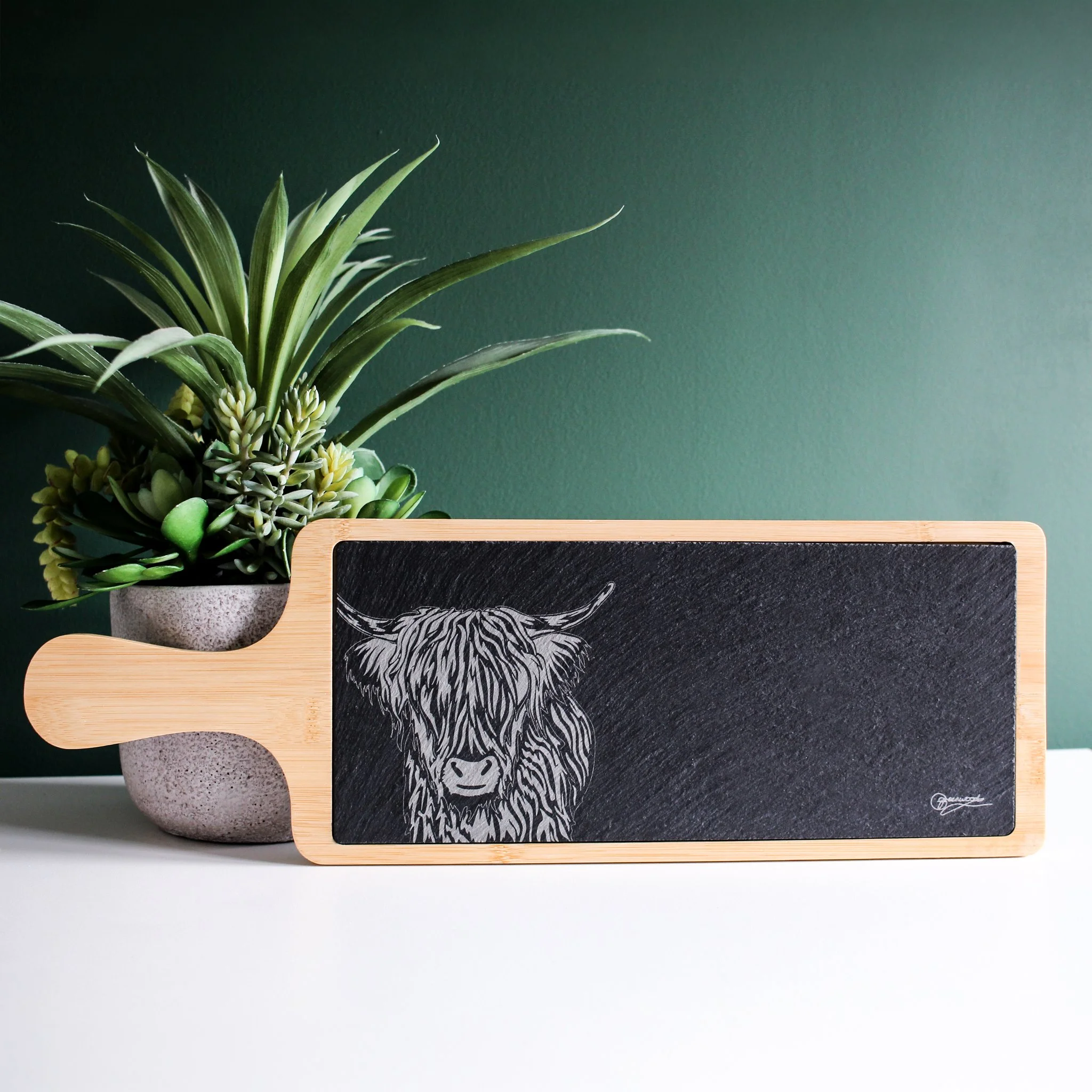 Highland cow bamboo and slate serving platter, displayed upright against plant pot and dark green background.