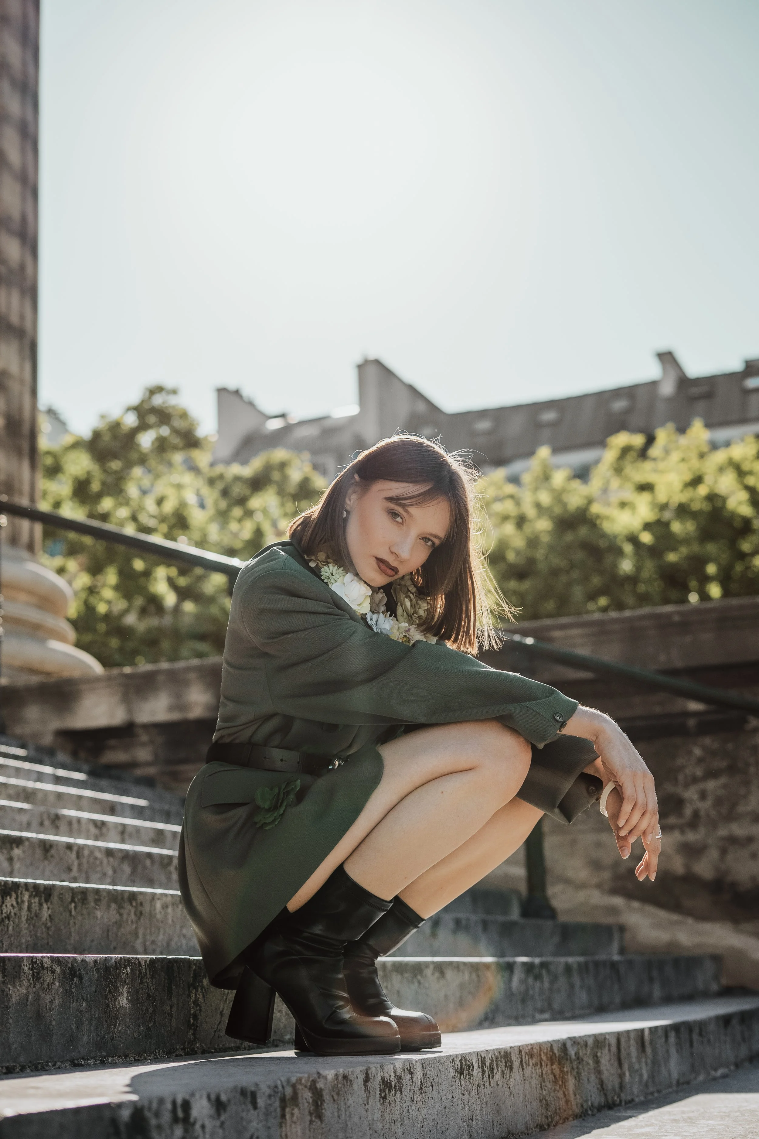 Jeune femme en vêtements verts, portant des bottes noires, assise sur des marches extérieures avec des arbres et bâtiments en arrière-plan, éclairée par le soleil.