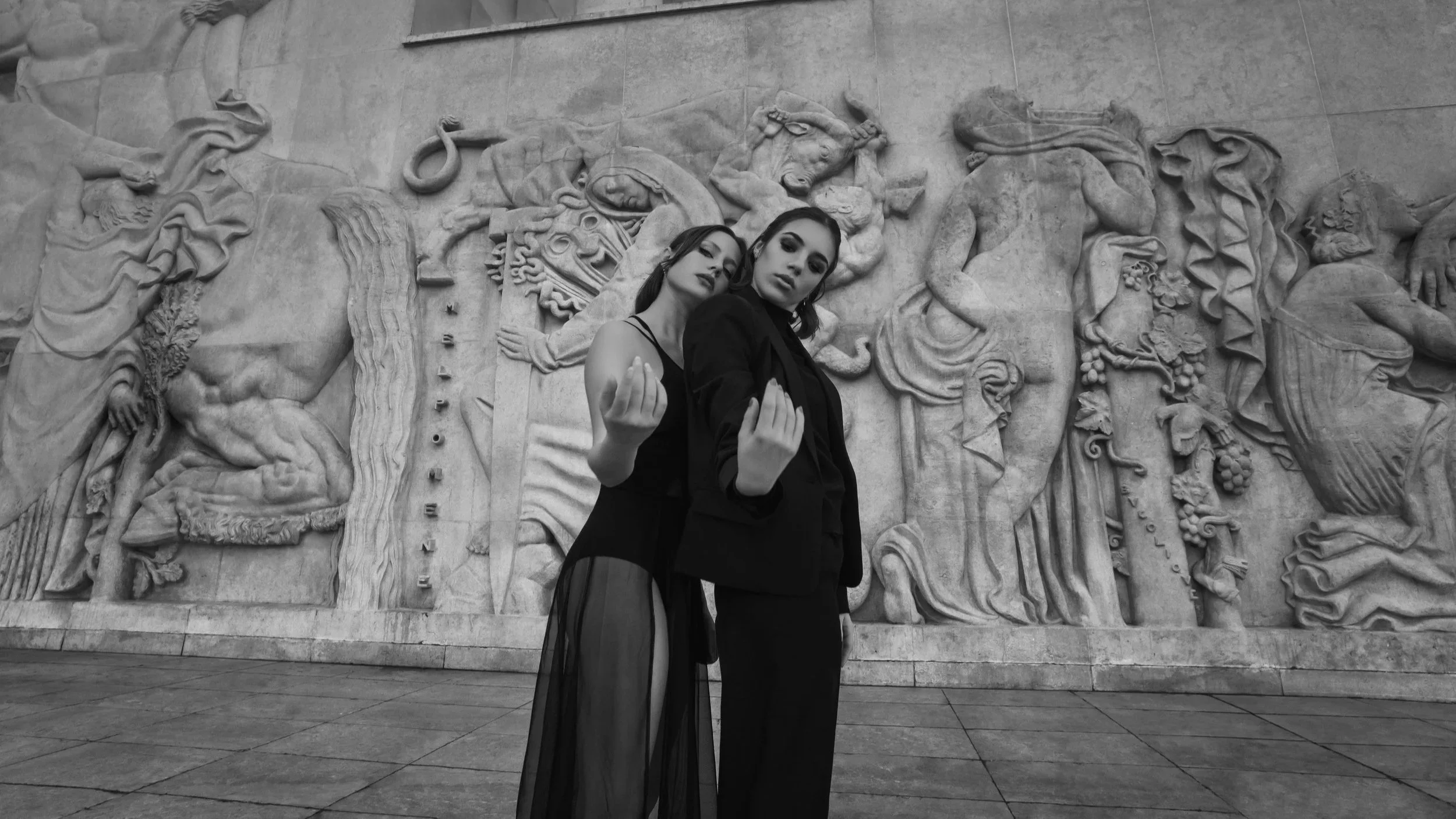 Deux femmes en vêtements noirs posant devant une sculpture en relief en pierre, avec des figures mythologiques ou historiques, dans un contexte urbain.