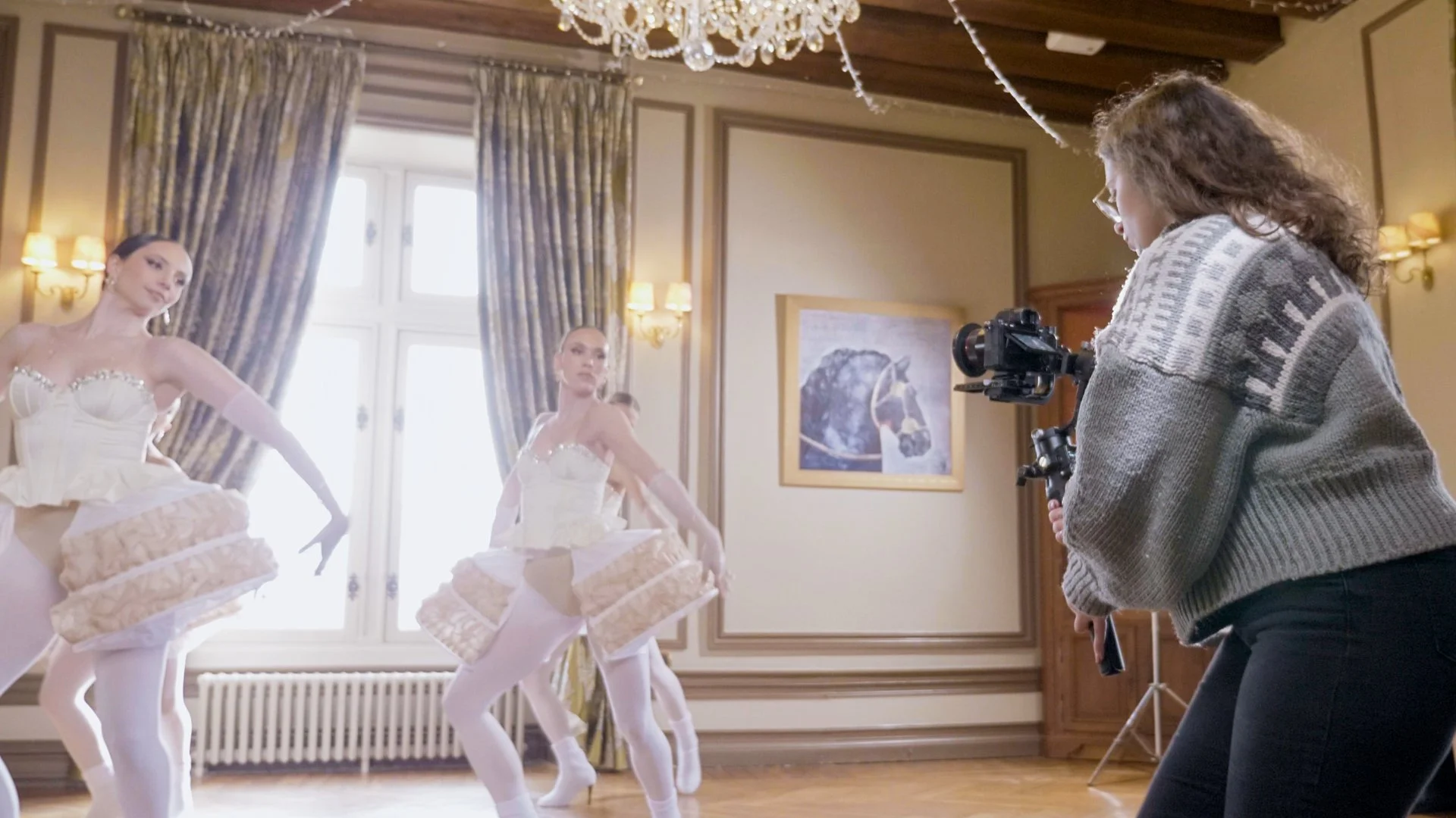 Une femme filme trois ballerines qui dansent dans une salle élégante avec de grandes fenêtres et des rideaux lourds.