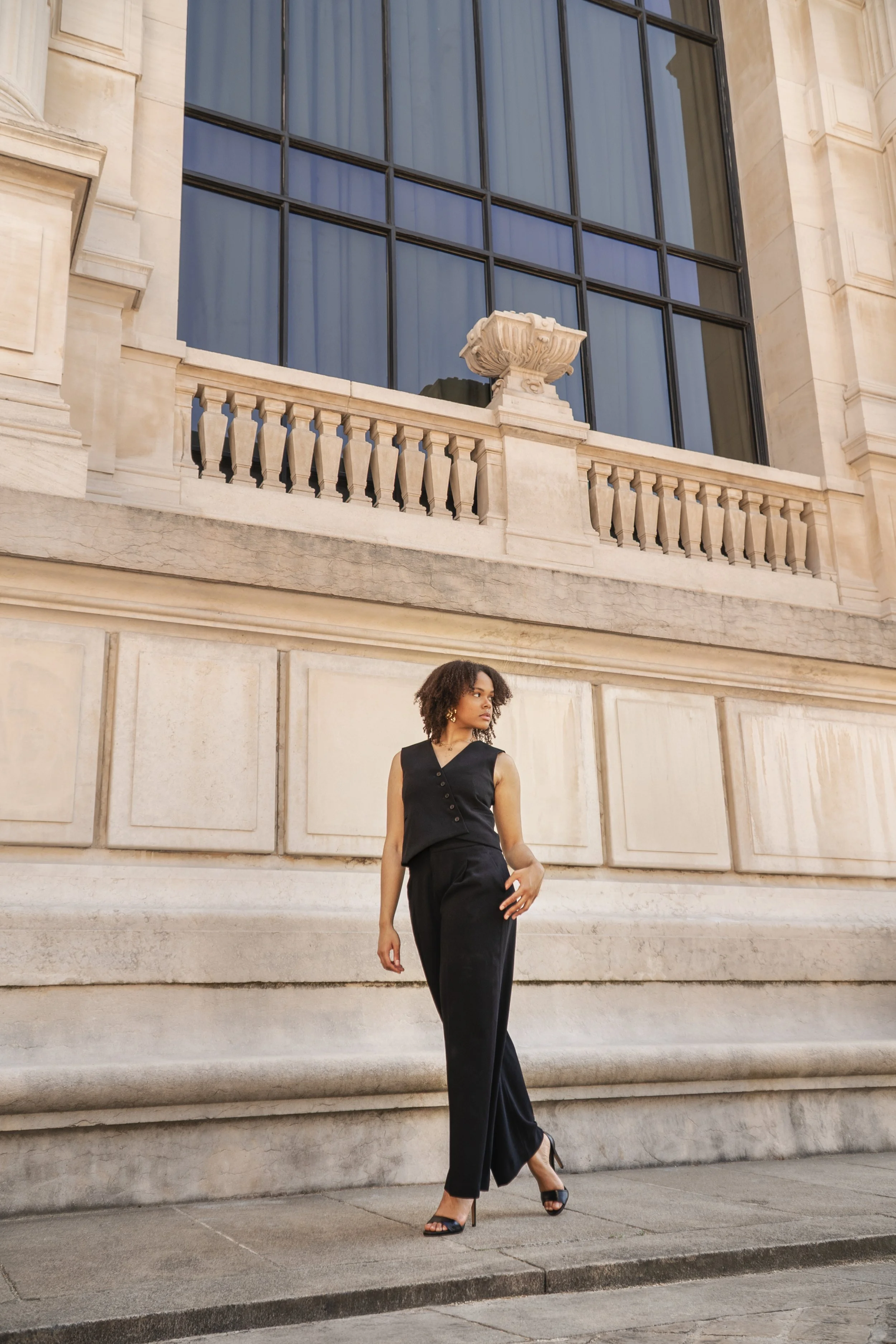 Jeune femme élégante portant une combinaison noire, debout devant un bâtiment en pierre avec de grandes fenêtres en verre et une balustrade décorative, en milieu urbain.