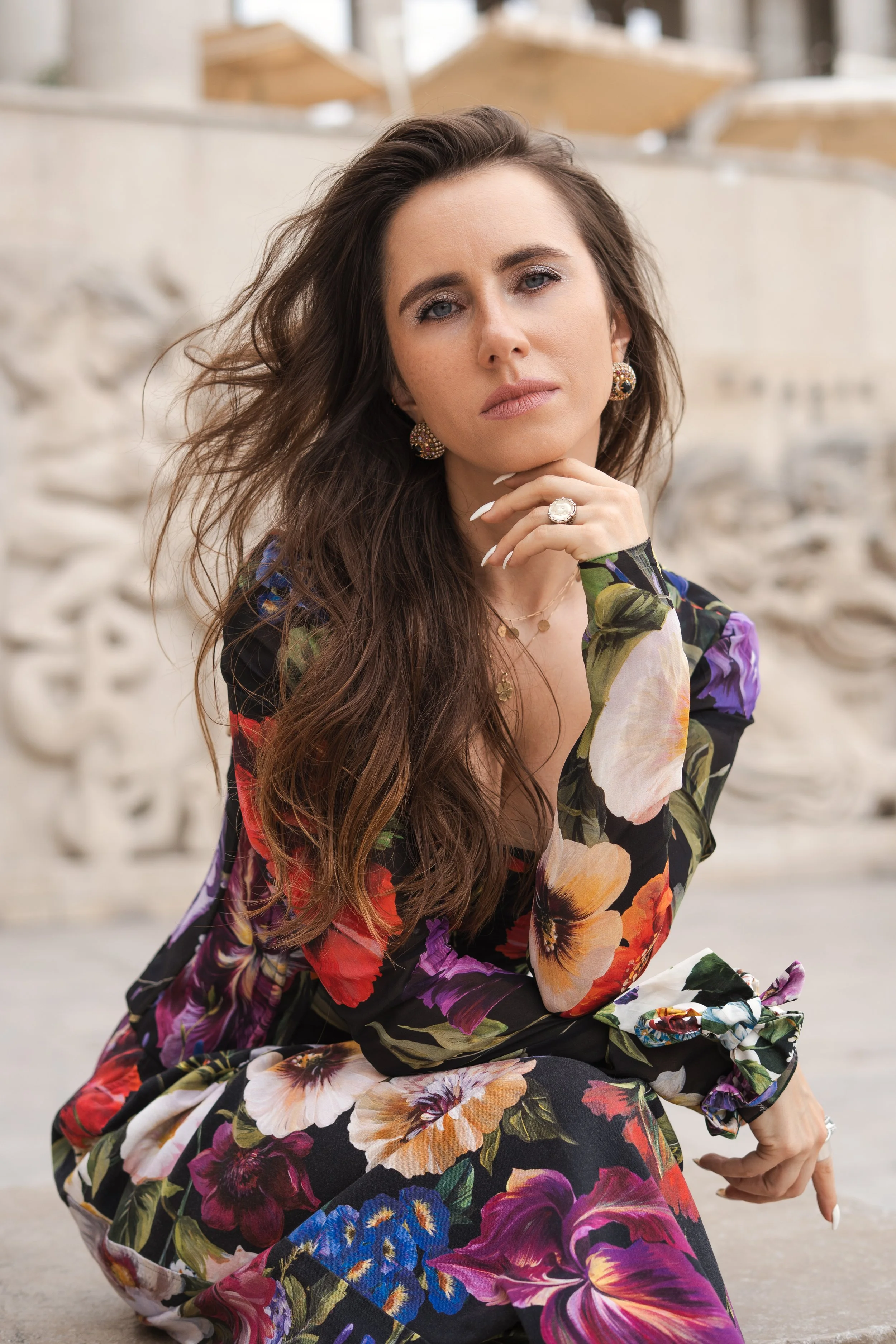 Femme avec longs cheveux bruns, portant des boucles d'oreilles, une bague et un collier, habillée d'un ensemble floral, assise devant un décor en pierre.