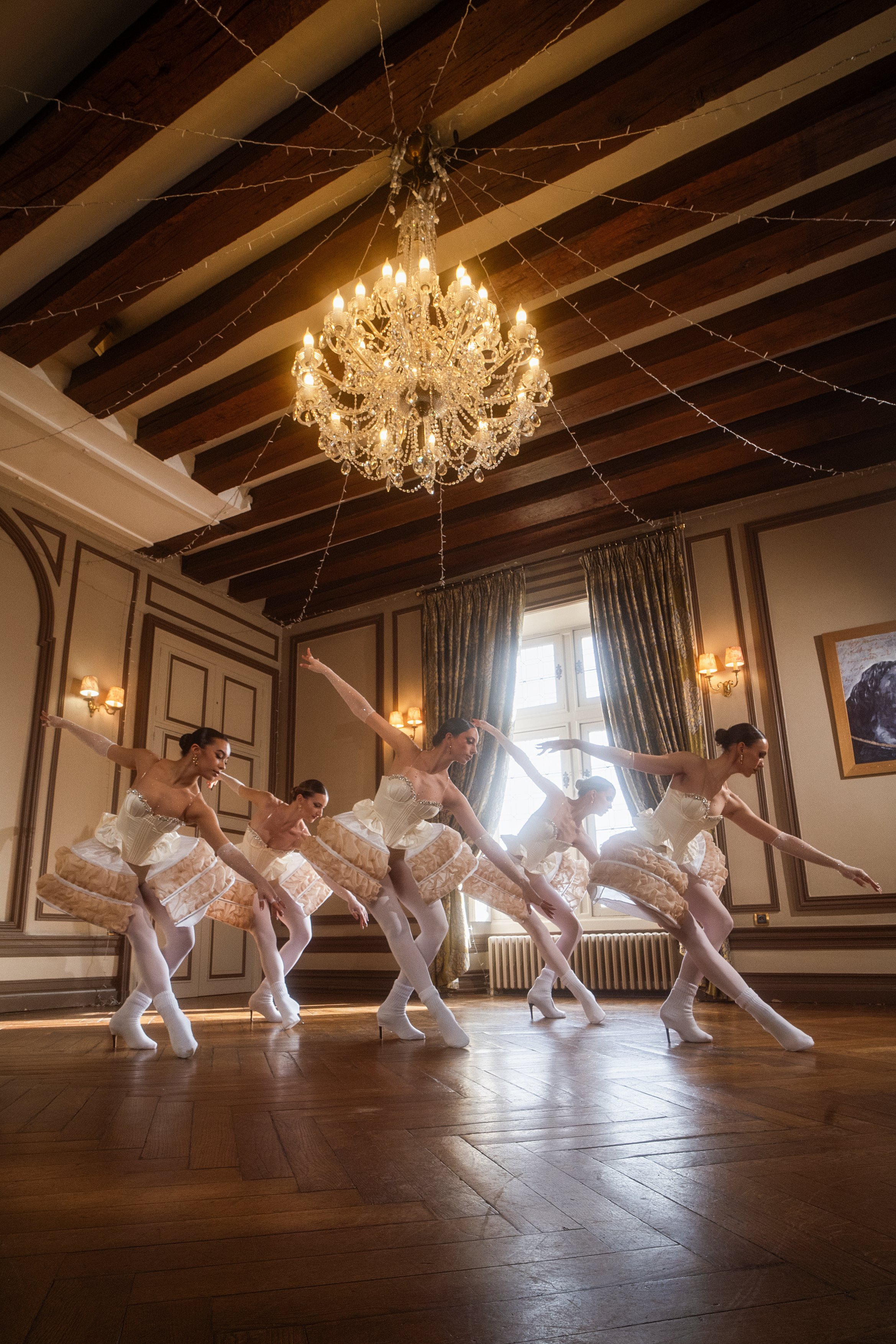 Cinq ballerines en tutu blanc pratiquent la danse dans un salon élégant avec plafonds en bois, lustre en cristal et grande fenêtre avec rideaux dorés.