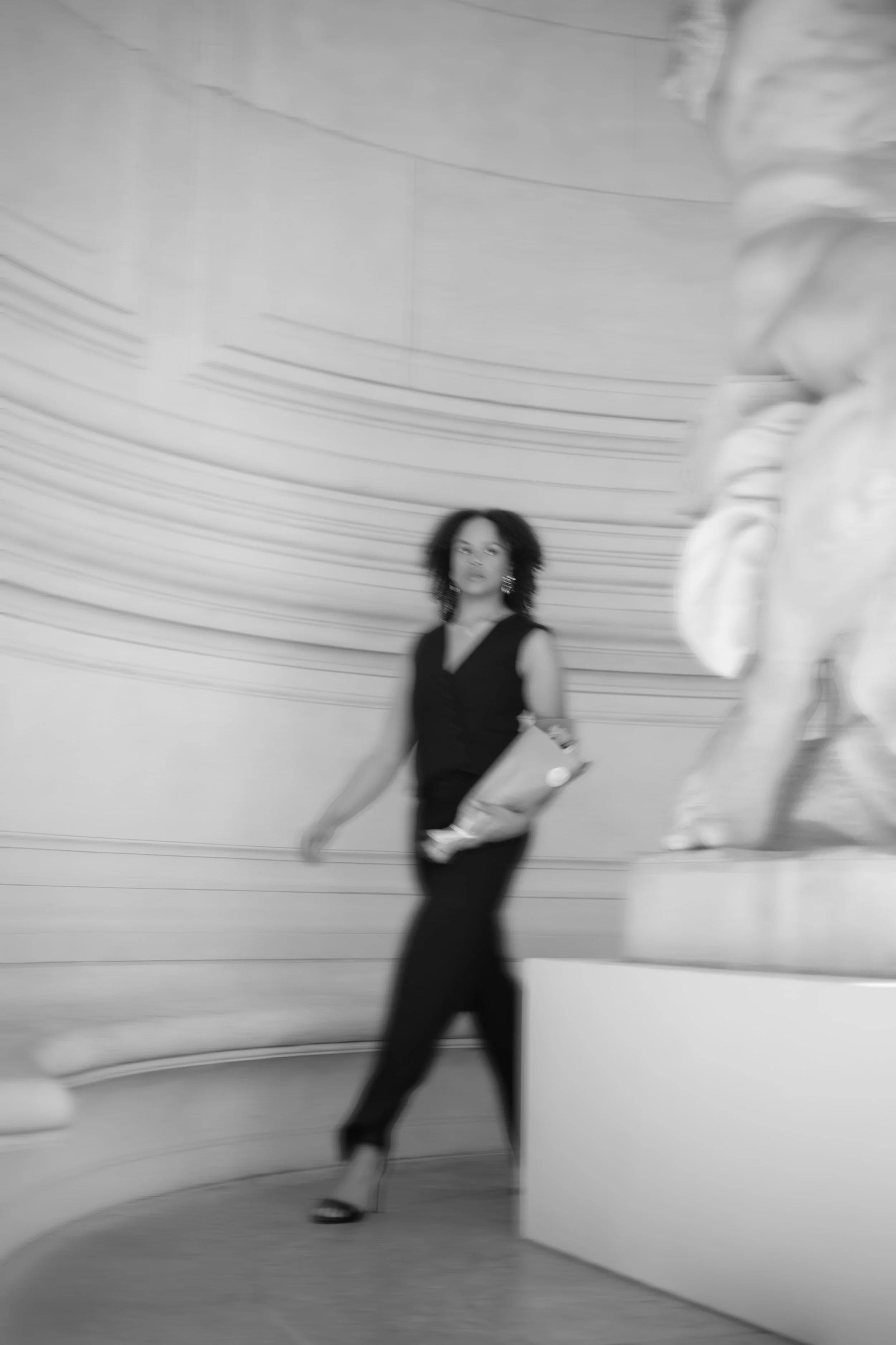 Une femme portant des vêtements noirs, portant un bouquet de fleur, se déplaçant dans une galerie avec une sculpture visible sur le côté droit.