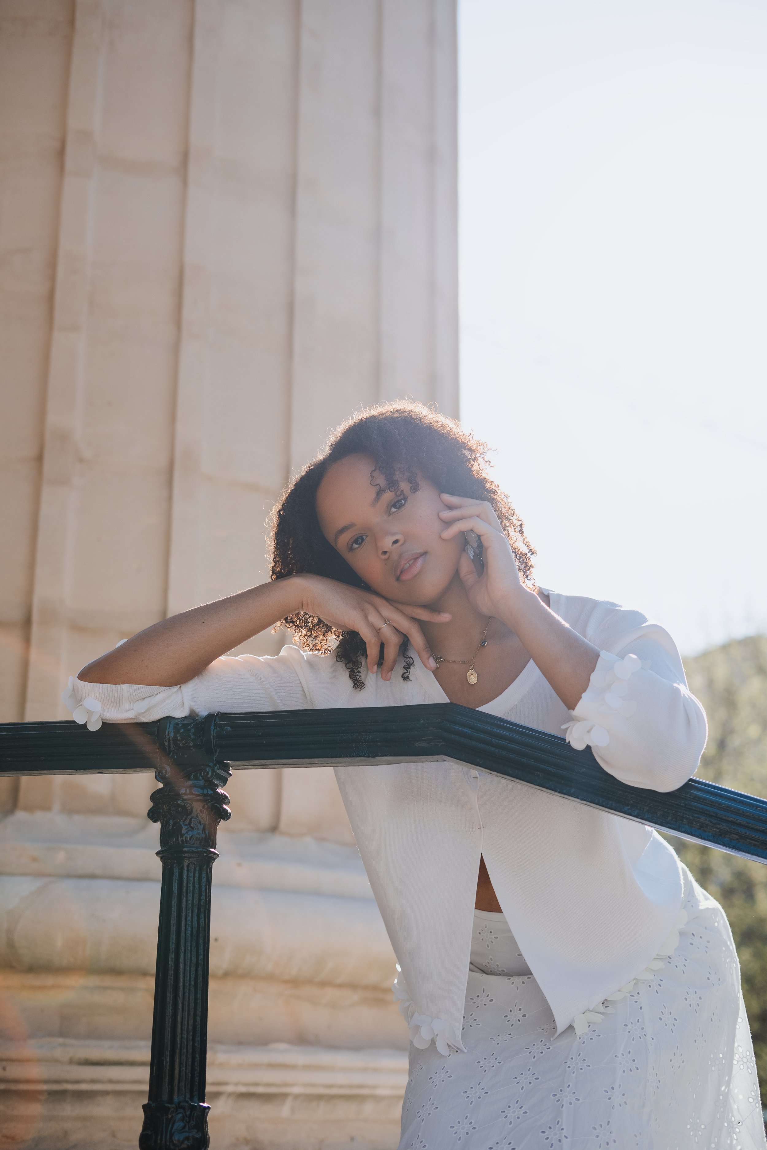 Jeune femme posant près d'une structure en pierre avec un rayon de lumière en arrière-plan, portant une robe blanche avec des motifs brodés et un cardigan blanc.