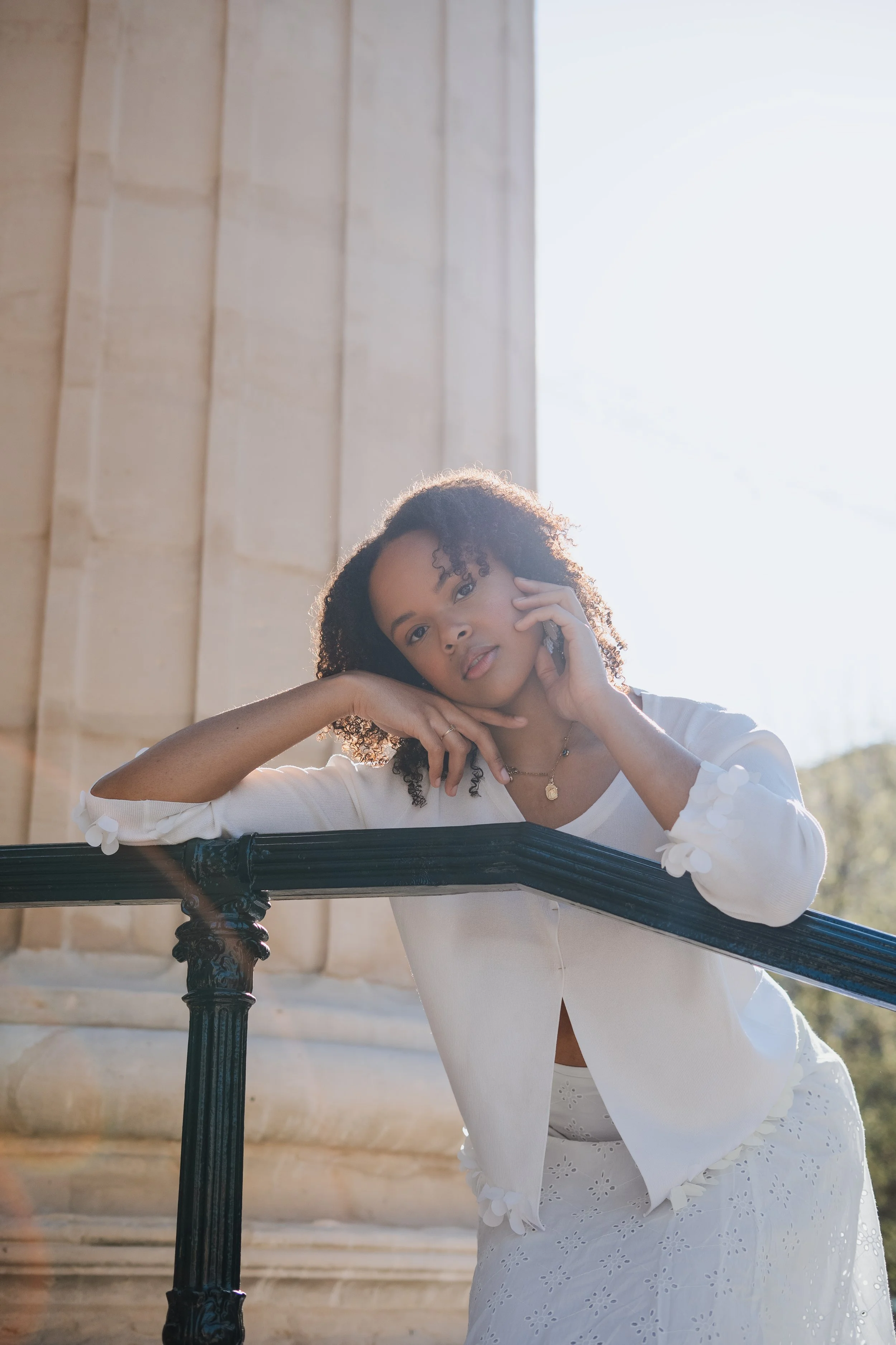 Jeune femme noire posant au soleil, portant une blouse blanche et une jupe en dentelle, avec un bâtiment en pierre en arrière-plan