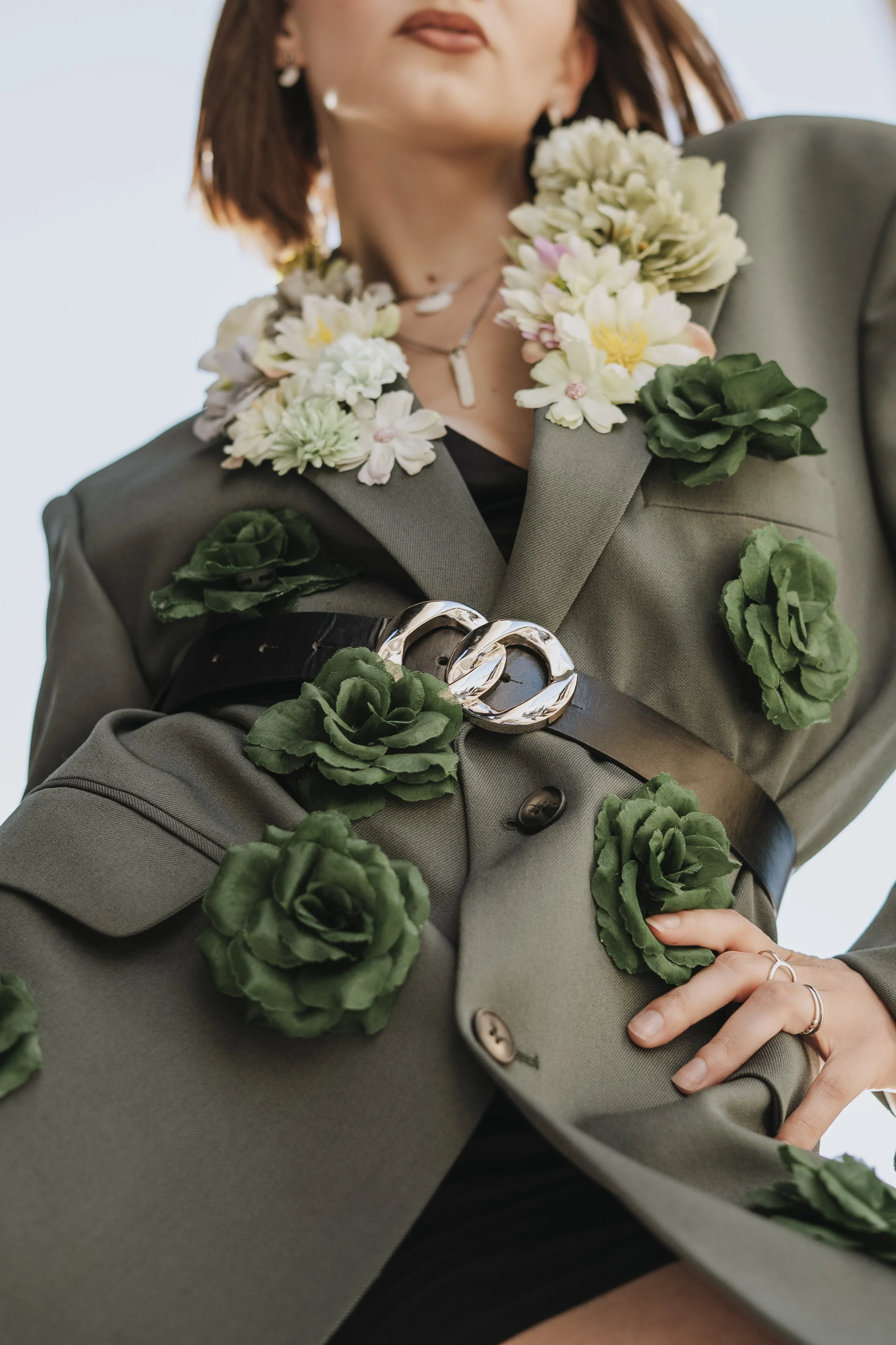 Une femme portant un costume vert décoré de fleurs en tissu vert et blanc, avec des accessoires en argent et un porte-bagages noir.