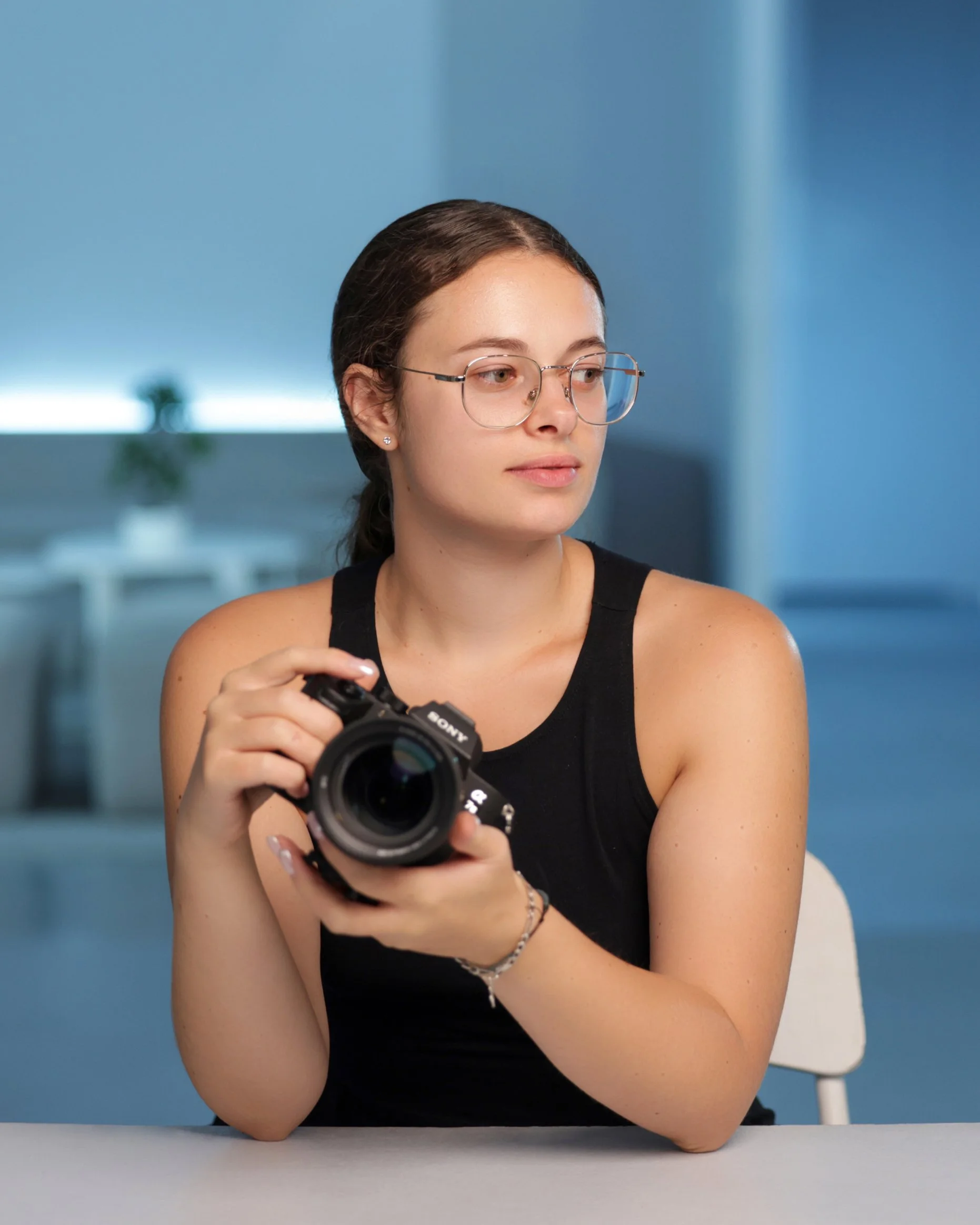 Jeune femme avec lunettes, portant un débardeur noir, tenant un appareil photo Sony dans les mains, assise à une table dans un intérieur moderne.