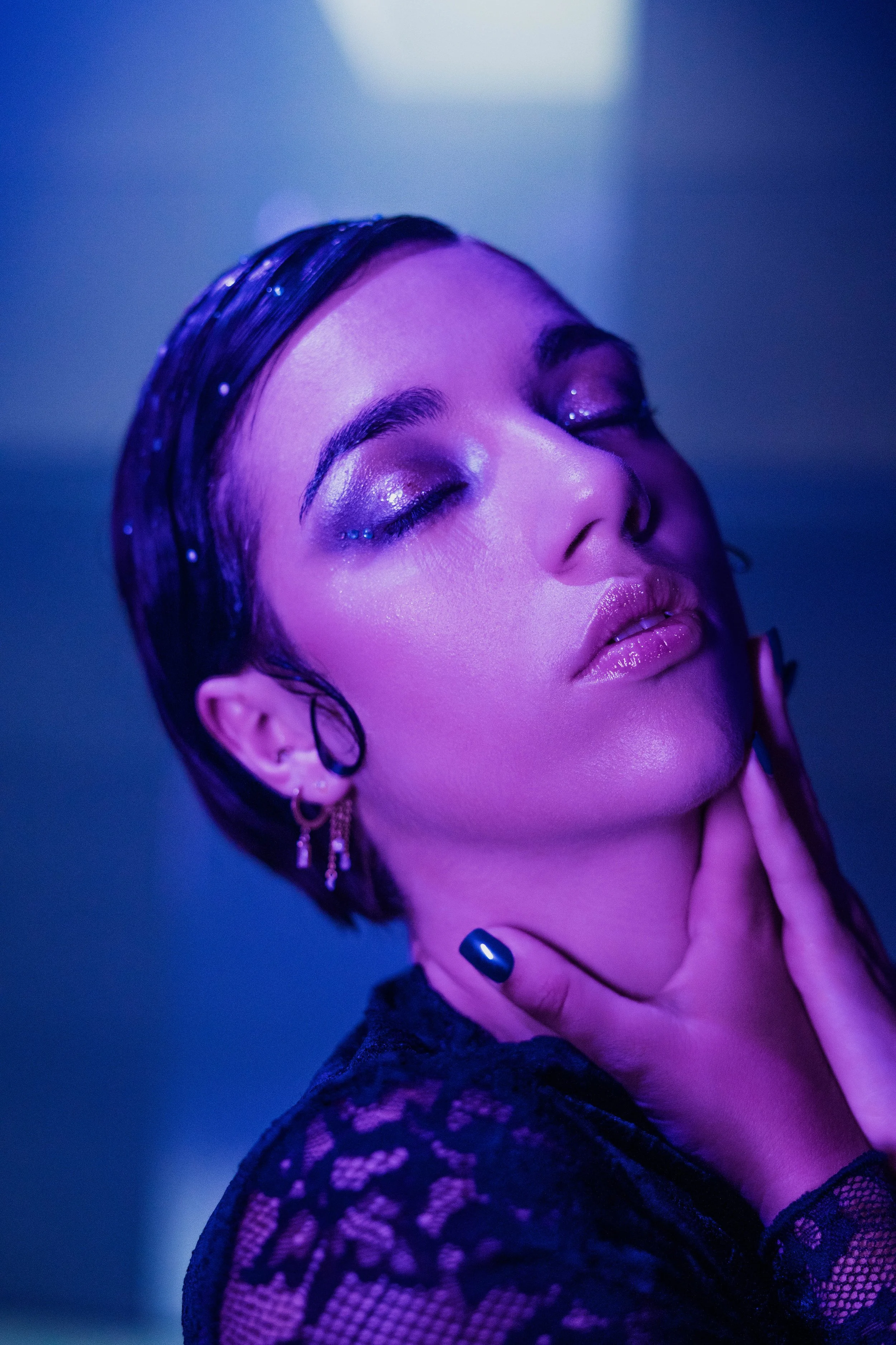 Une femme avec des cheveux courts, maquillage scintillant et lèvres brillantes, touche son cou dans un éclairage violet.