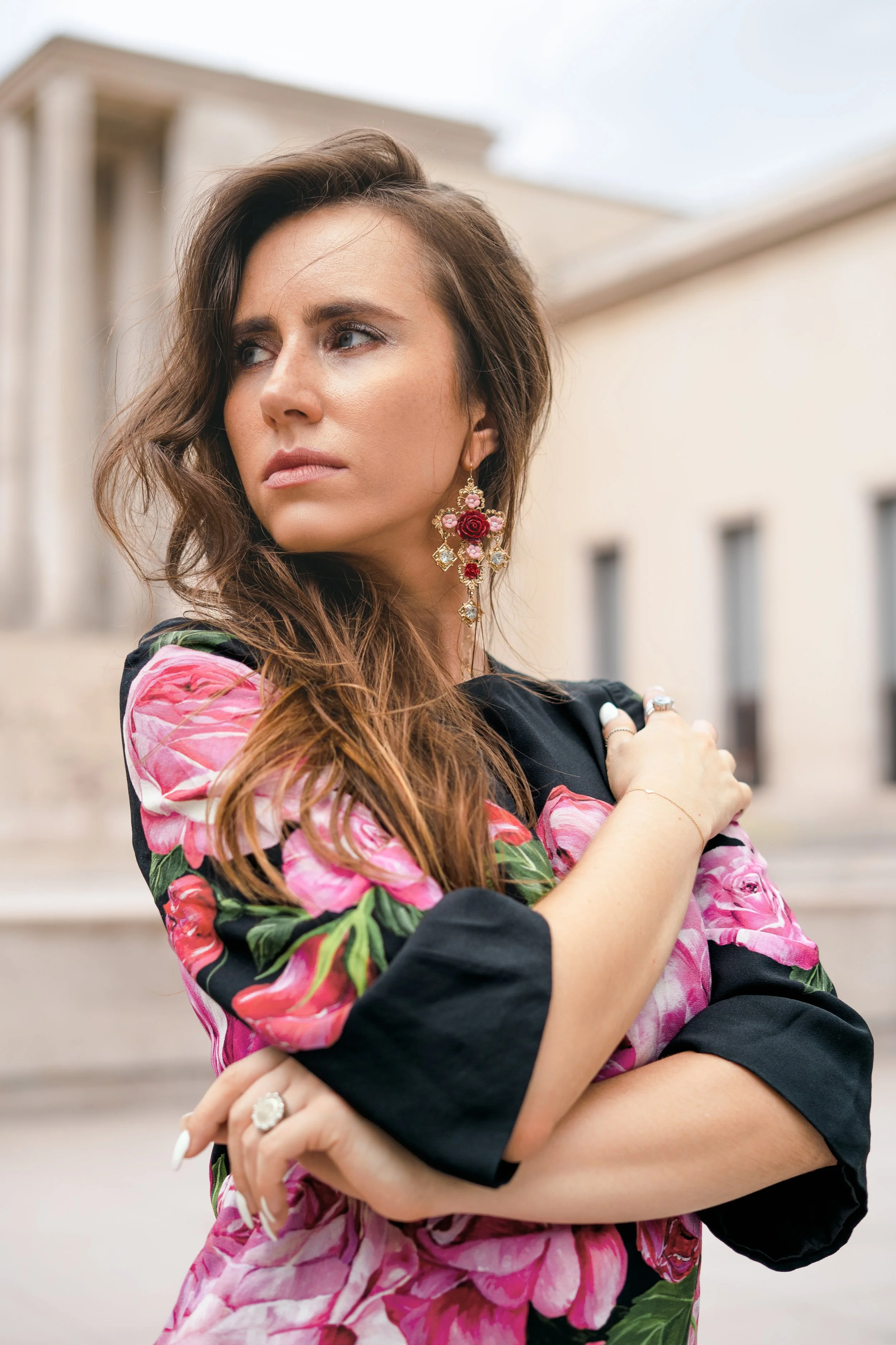 Femme avec longs cheveux bruns portant une robe florale noire et rose, portant des boucles d'oreilles complexes et des bijoux, avec un fond architectural classique.