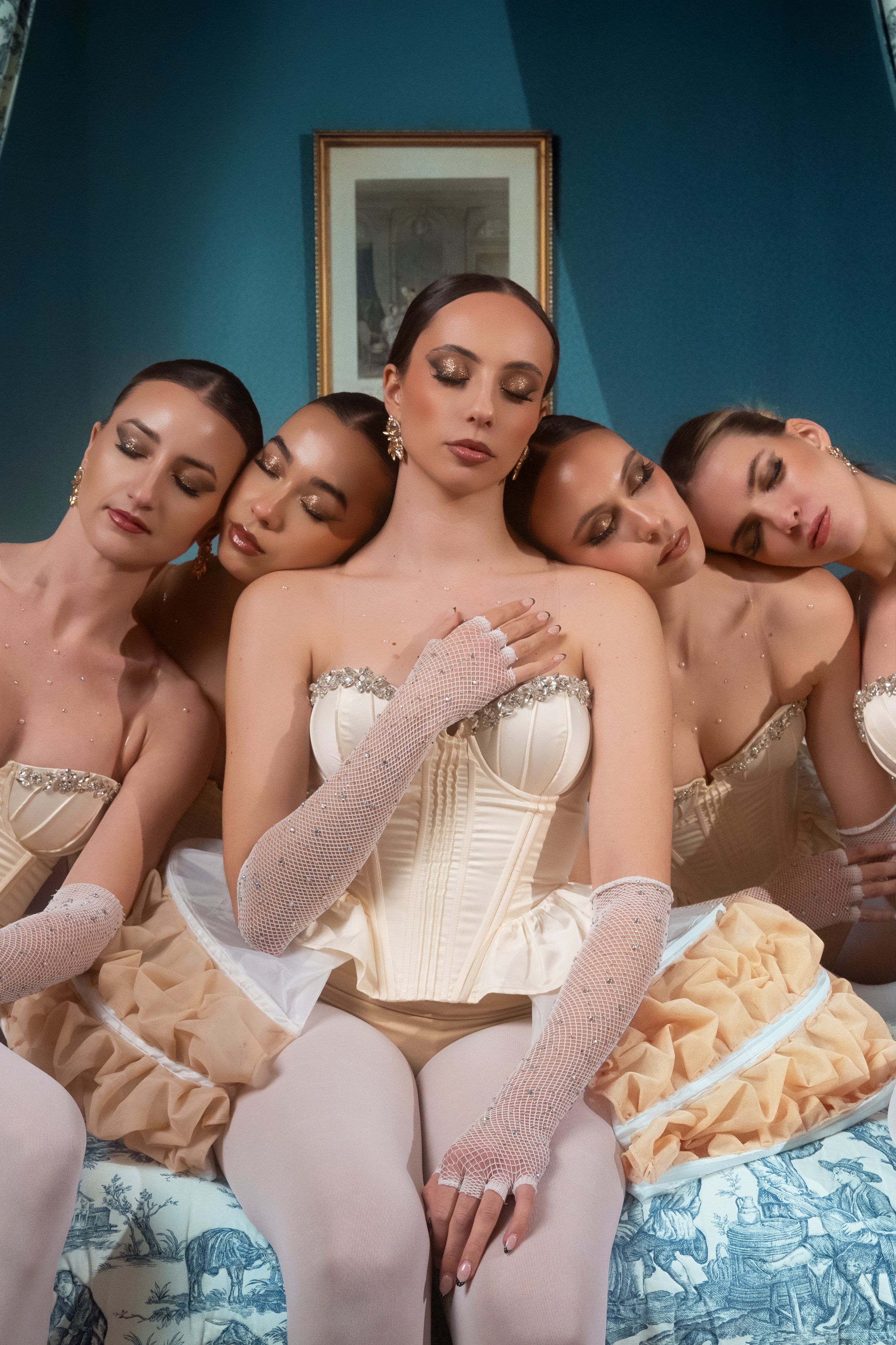 Cinq femmes assises ensemble, portant des corsets beige, des gants en maille blanche, des jupes en tissu beige et des collants blancs, avec un fond bleu et un tableau encadré derrière elles.