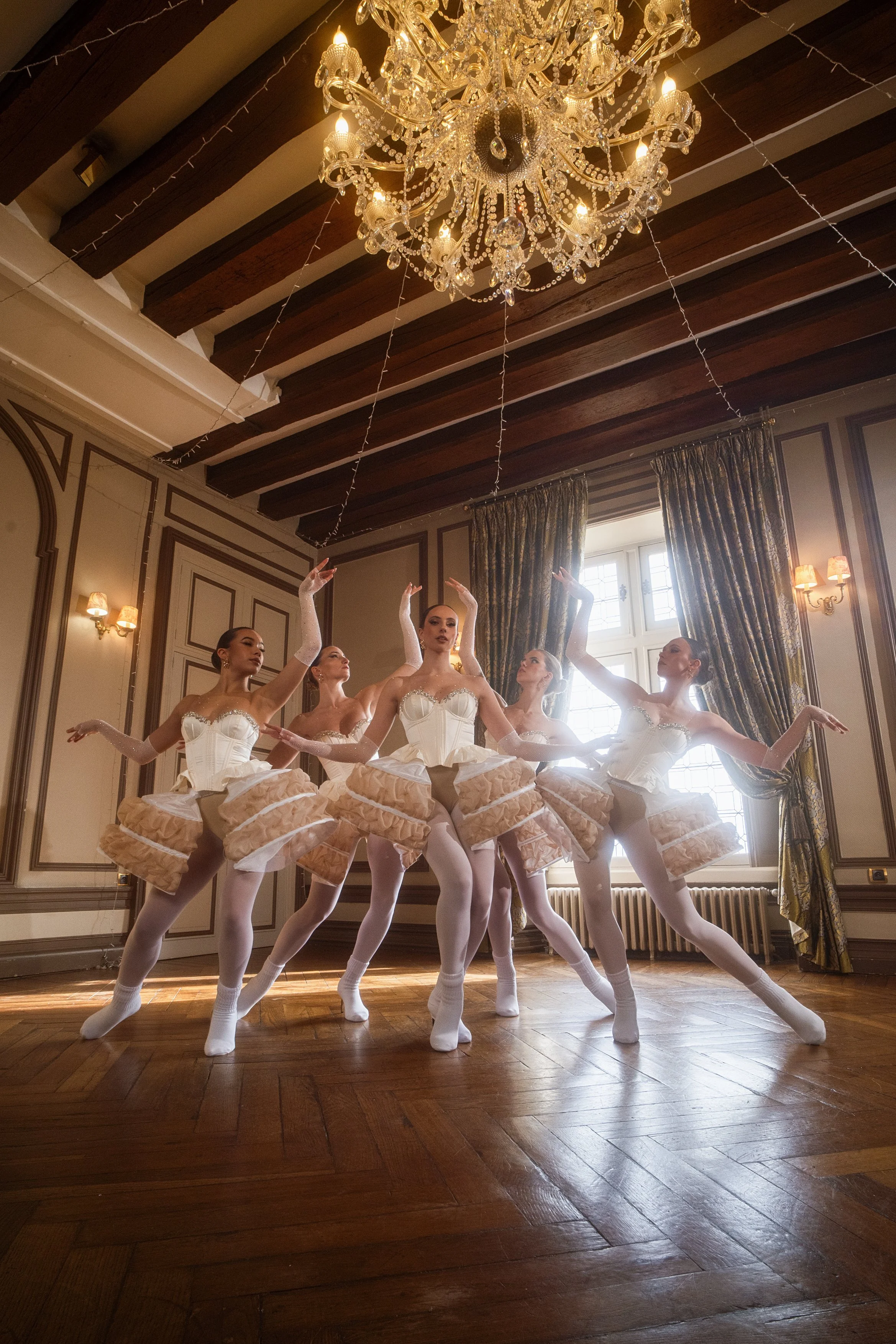 Cinq danseuses en tutu beige, posant dans une salle élégante avec lustre en cristal et grandes fenêtres.