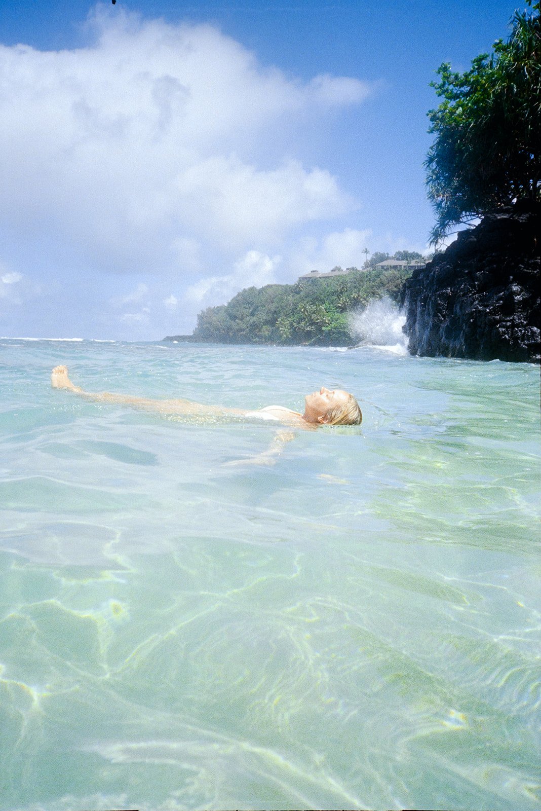 A person with blonde hair floating peacefully on their back in clear, shallow ocean water near a rocky shoreline with lush green trees and a blue sky.