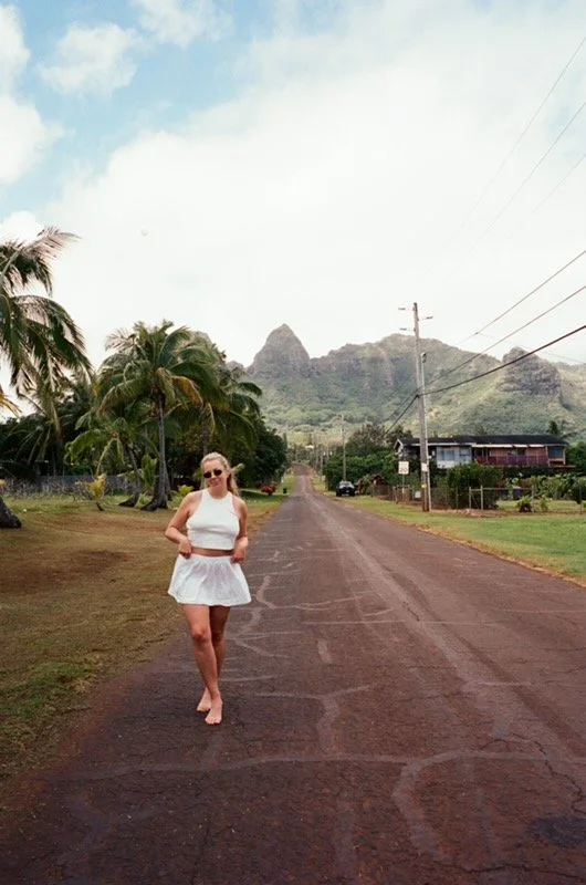 A woman in a white skirt and crop top walking barefoot on a rural road with palm trees and mountains in the background.