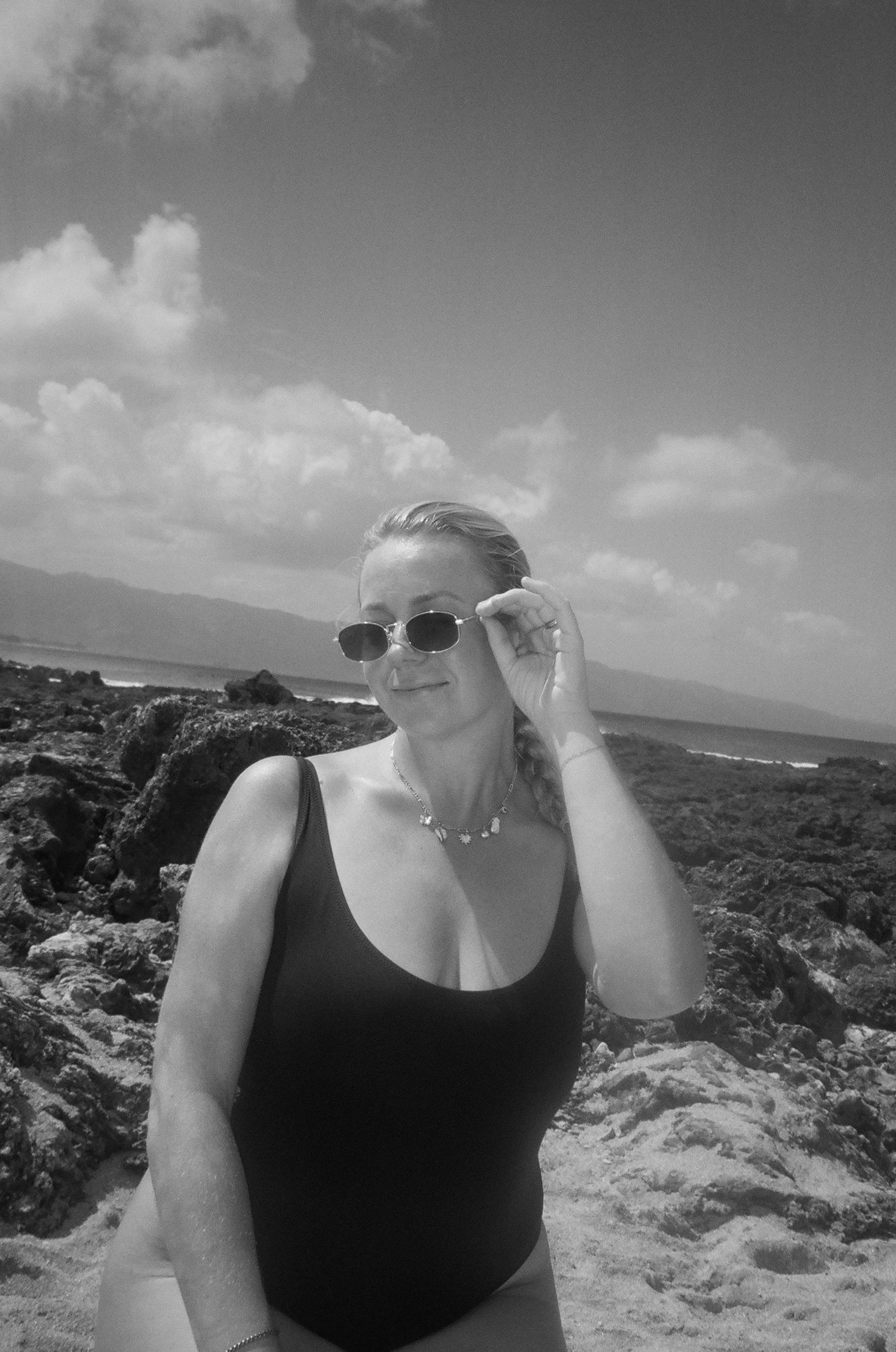 A woman with sunglasses and a necklace at the beach, with rocky shoreline and mountains in the background, under a partly cloudy sky.
