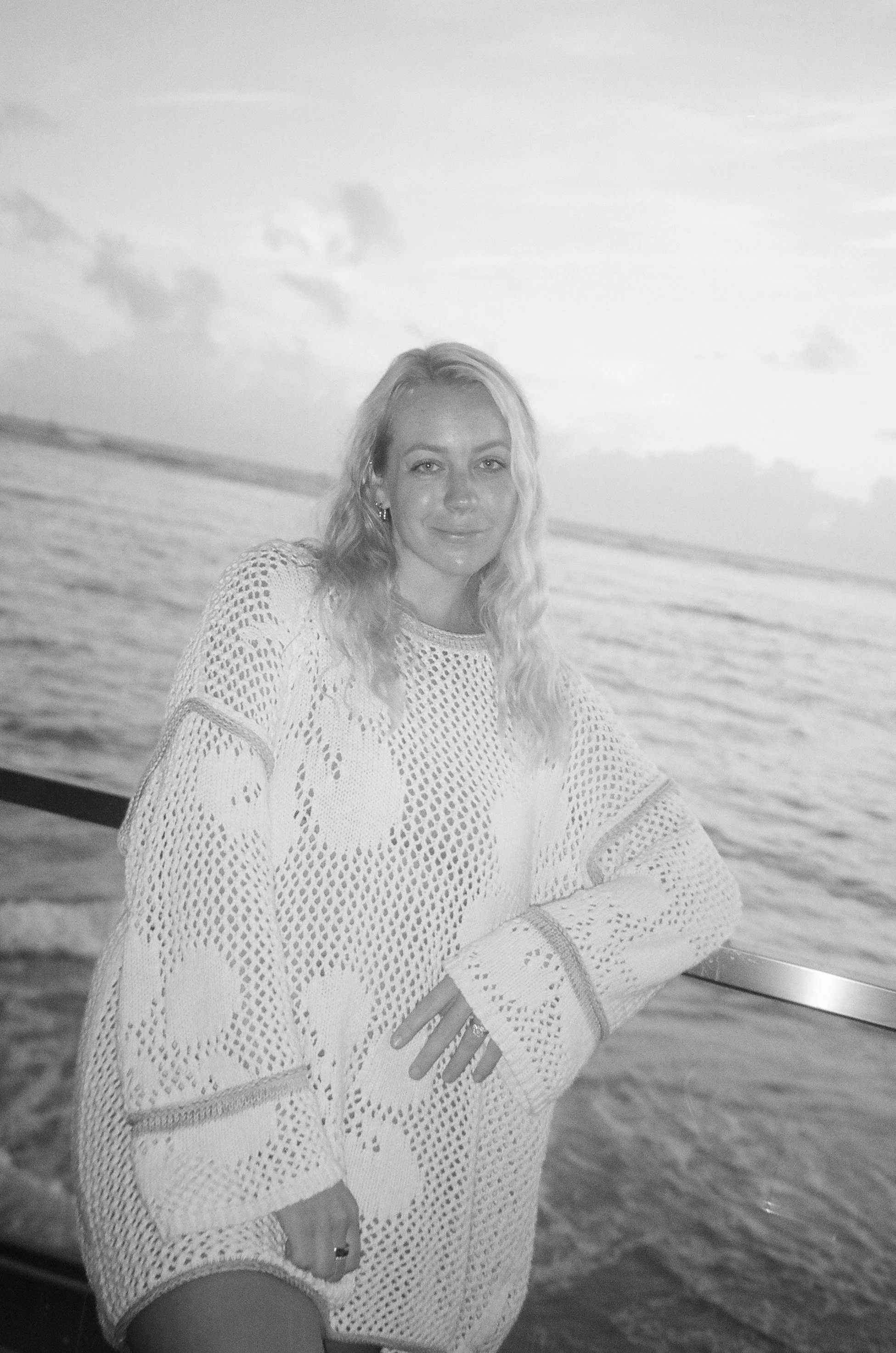 Woman with long wavy hair in a crochet sweater, standing by the ocean at sunset, with clouds in the sky.