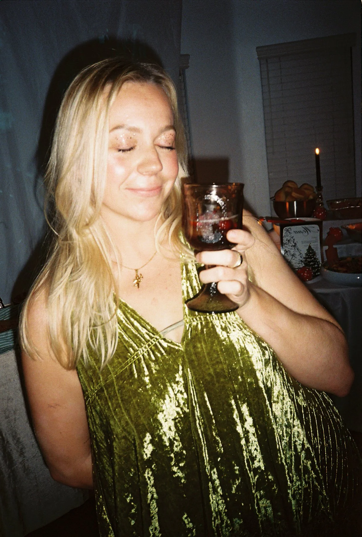 A woman with blonde hair in a green velvet dress holding a glass of red wine, smiling with her eyes closed at a festive gathering.