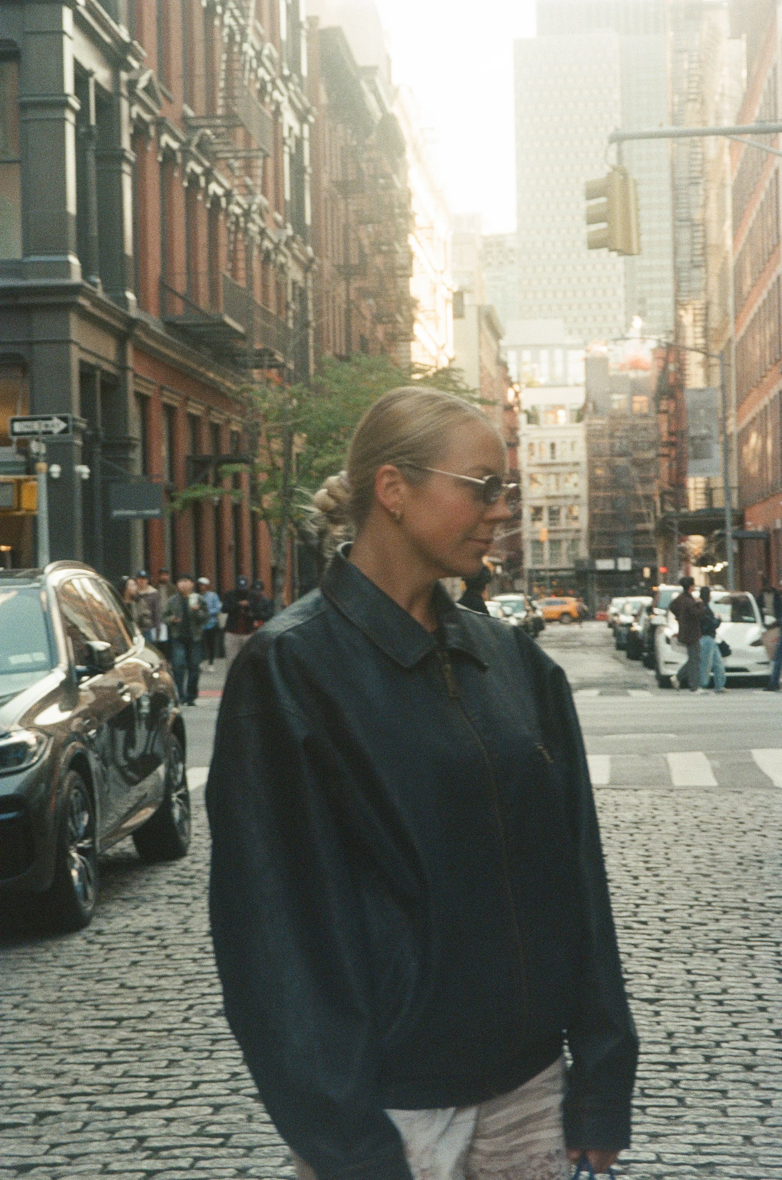A woman with blonde hair tied back, wearing sunglasses and a dark jacket, standing on a city street with tall buildings, parked cars, and pedestrians in the background.