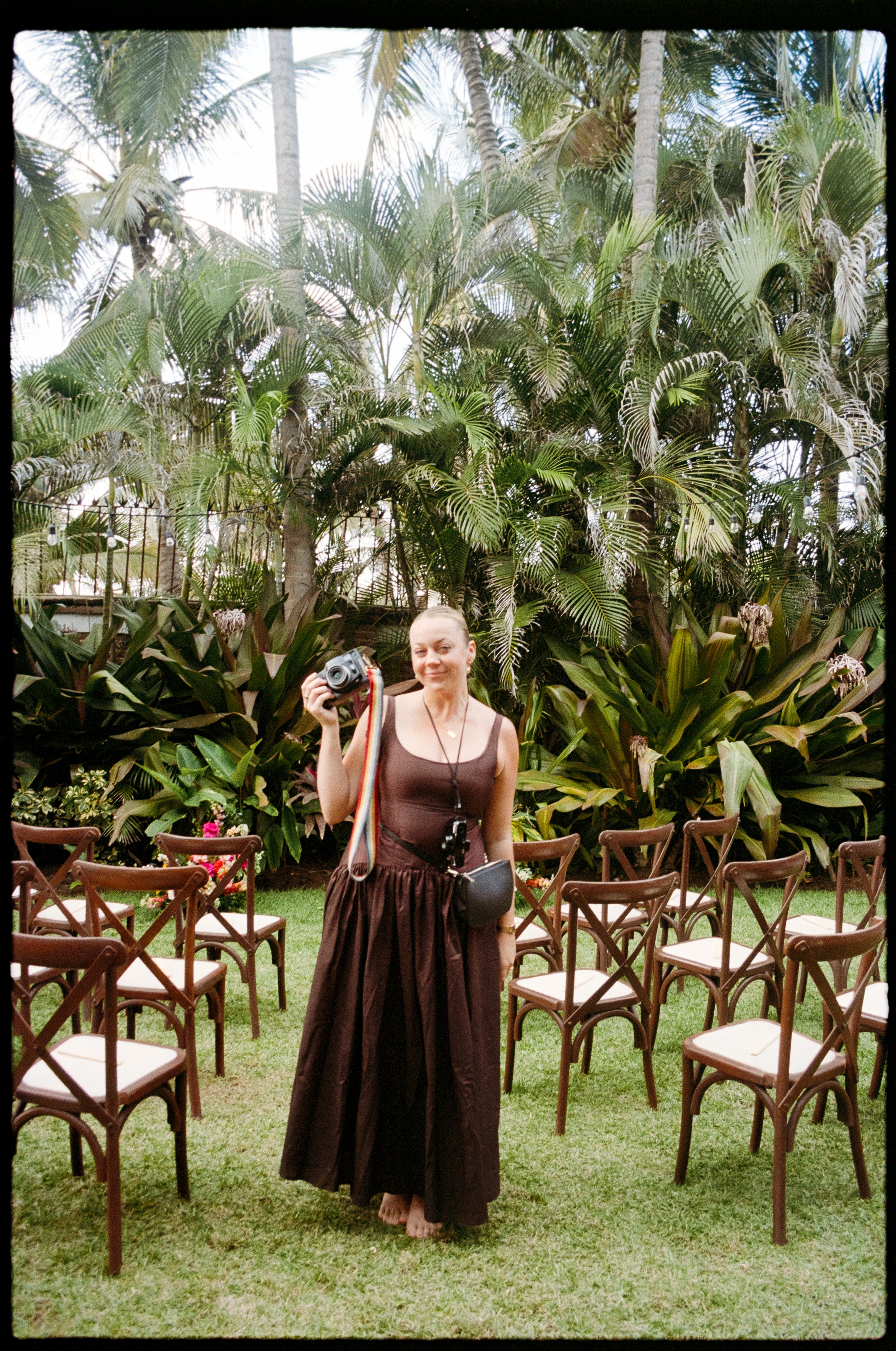 A young woman in a brown strapless top and a long brown skirt stands barefoot on grass, holding a camera. The background shows a lush, tropical garden with tall palm trees and leafy plants, and empty chairs arranged in rows, indicating an outdoor event setup.