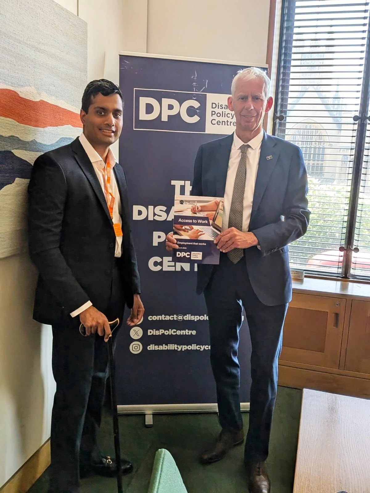 Arun from the Disability Policy Centre stands beside John Milne MP, who is holding the DPC’s Access to Work: Employment that Works report in front of a DPC banner.