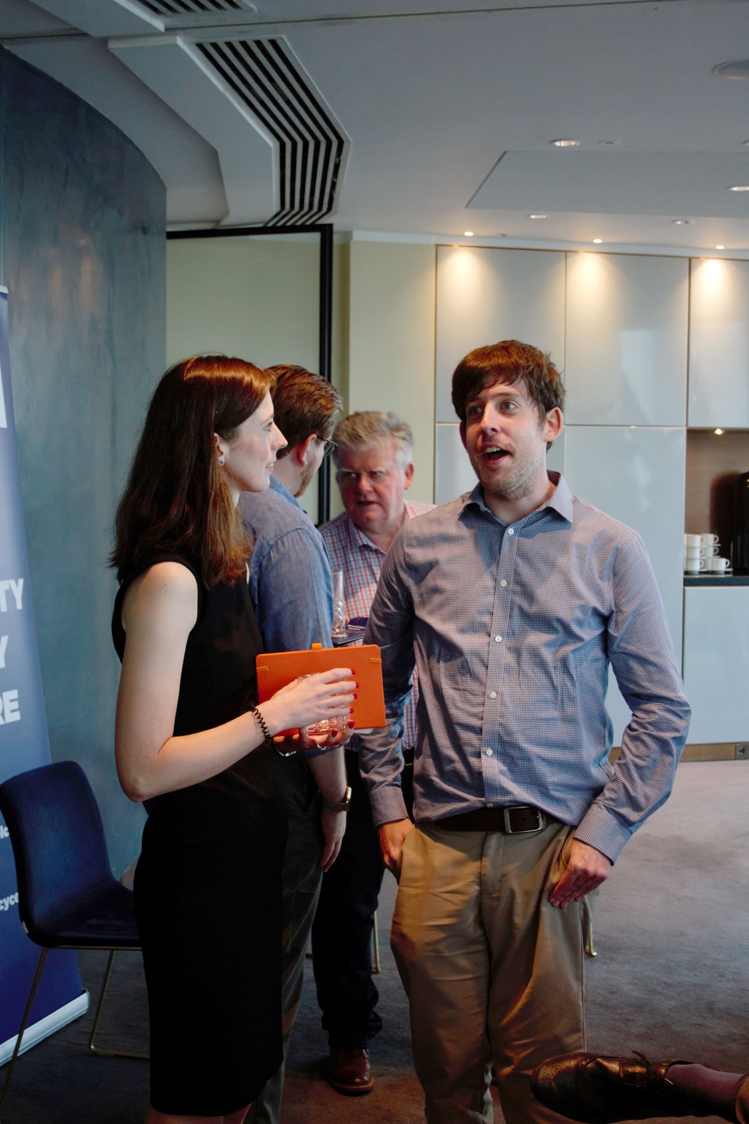 Photograph of Chloe and Mark stood up at the front of the room talking to each other. Chloe is to the left in a smart black dress holding a orange notebook. Mark on the right is dressed in light blue shirt and beige trousers.