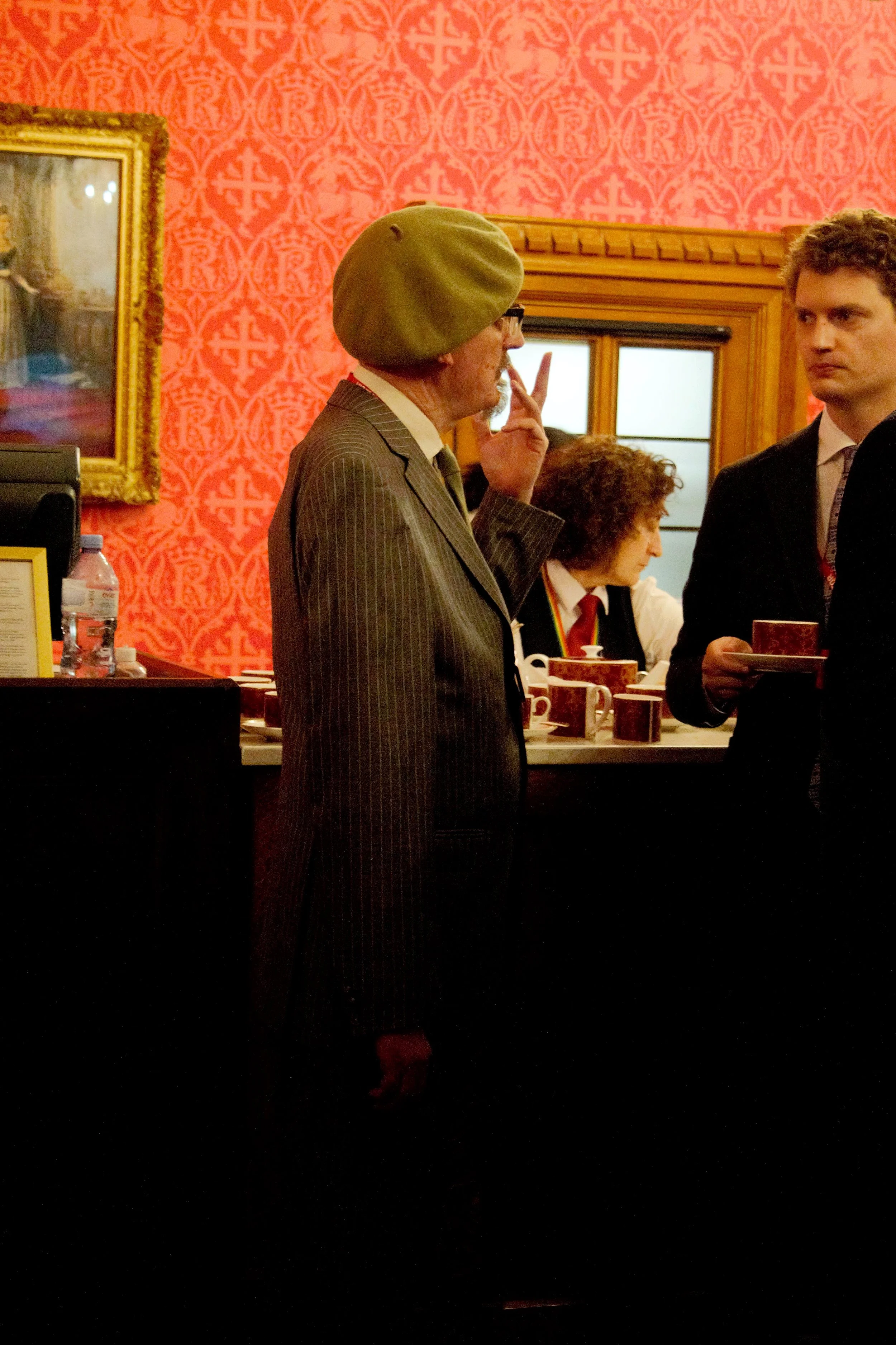 Side shot of a male guest speaking to another guest in the red room. He turned to the right, wearing a army green beret with a white shirt and a pin-stripped suit jacket 