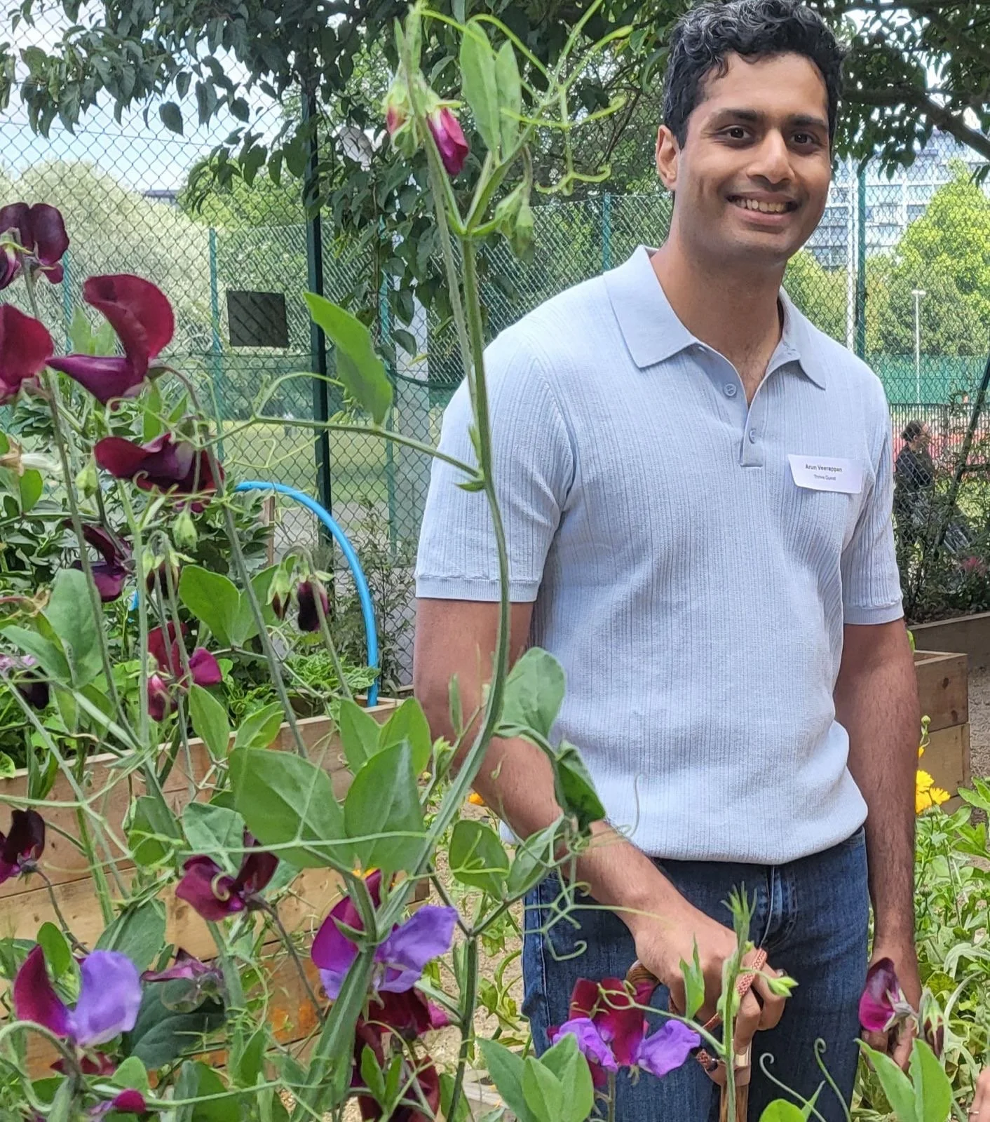 Arun Veerappan stands smiling in a community garden beside raised flower beds filled with purple sweet peas. He wears a light blue polo shirt and jeans, with trees and a fenced sports court visible behind him.
