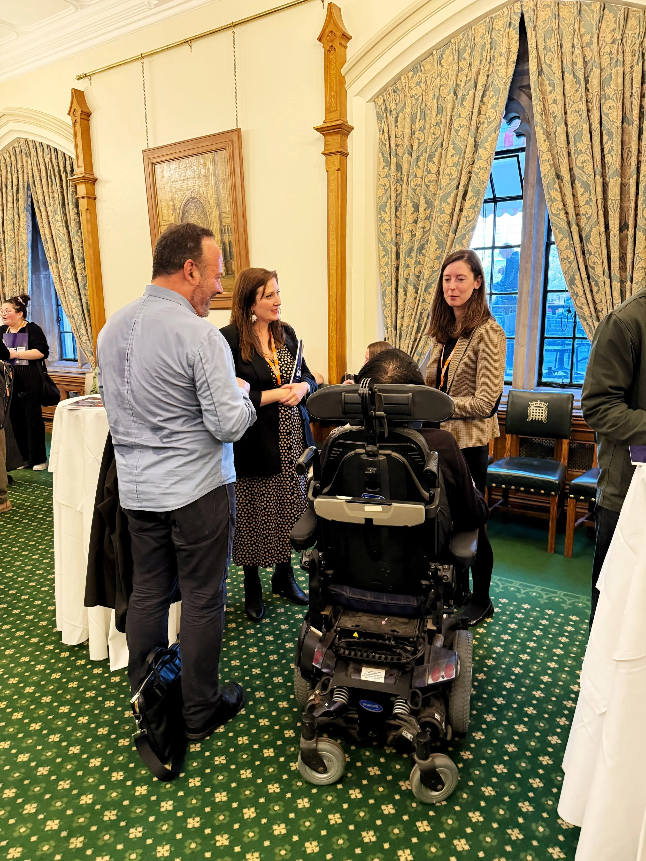 Conversation in the Churchill Room, Houses of Parliament, with three standing guest talking to wheelchair user. The room features chandeliers, green patterned carpet, tall curtains, and framed historic paintings on the walls.