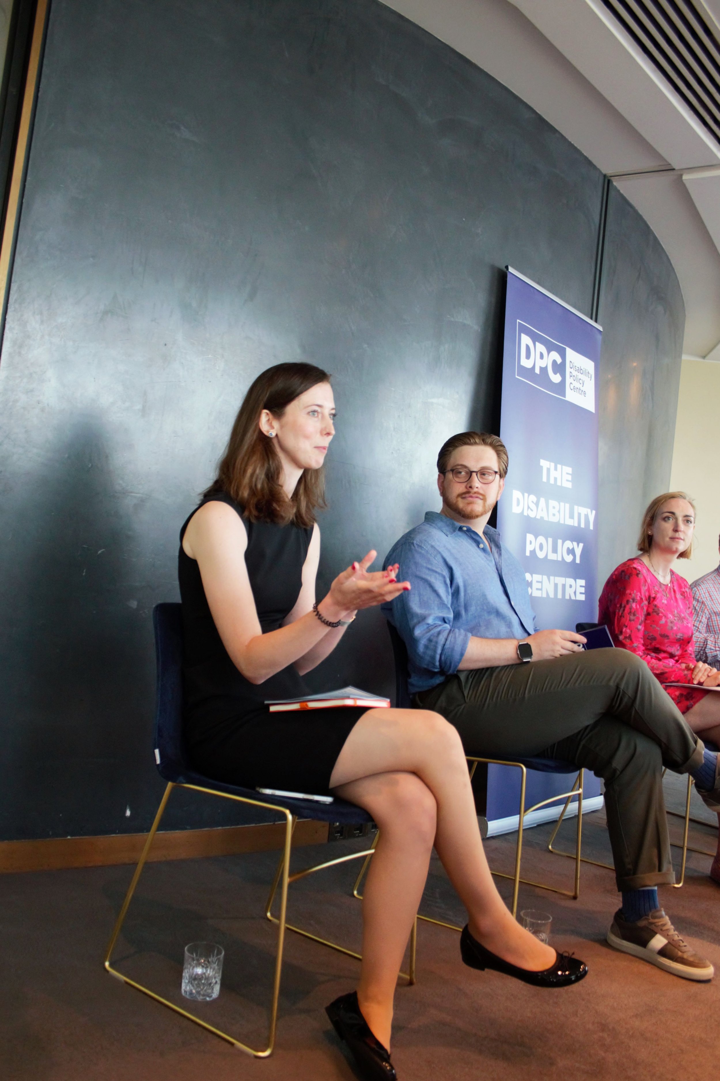 Photograph of chloe sat on a chair on the panel speaking. Next to her on her right is Joseph and Alys looking towards her. Chloe is in a smart balck dress and gesturing to the audience.