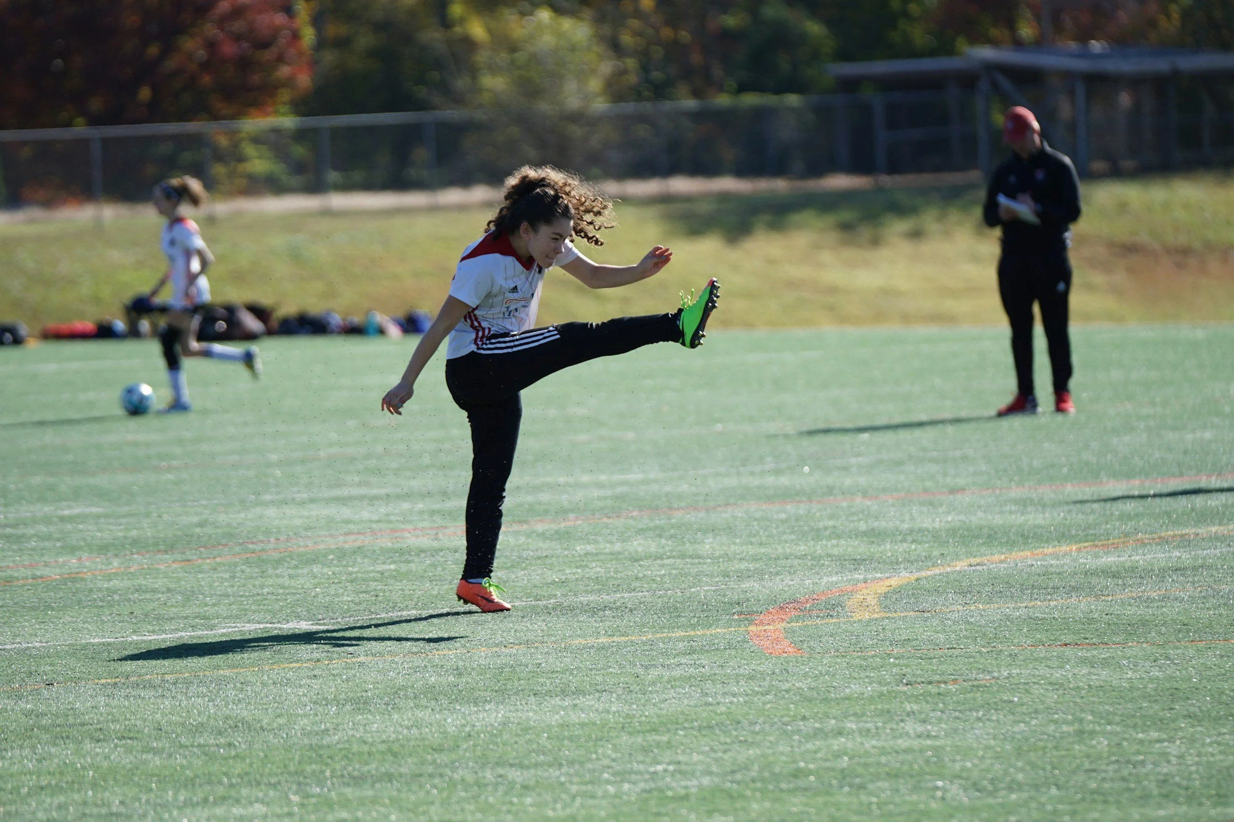 Photograph of young teenage girl on a football pitch kicking a ball with leg up in the air, she has long curly brown hair and white sports top, black tracksuit and green football boots on.