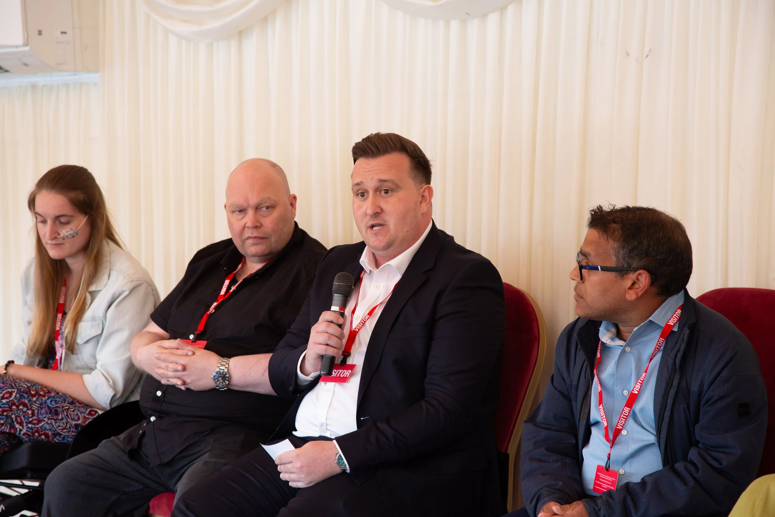 Close up of seated panel on the stage of guest speakers, starting from right , Dr Kush Kanodia, Daniel Cook, Matt Lloyd PLY and Mary FitzMeilton. Daniel cook is speaking with the microphone