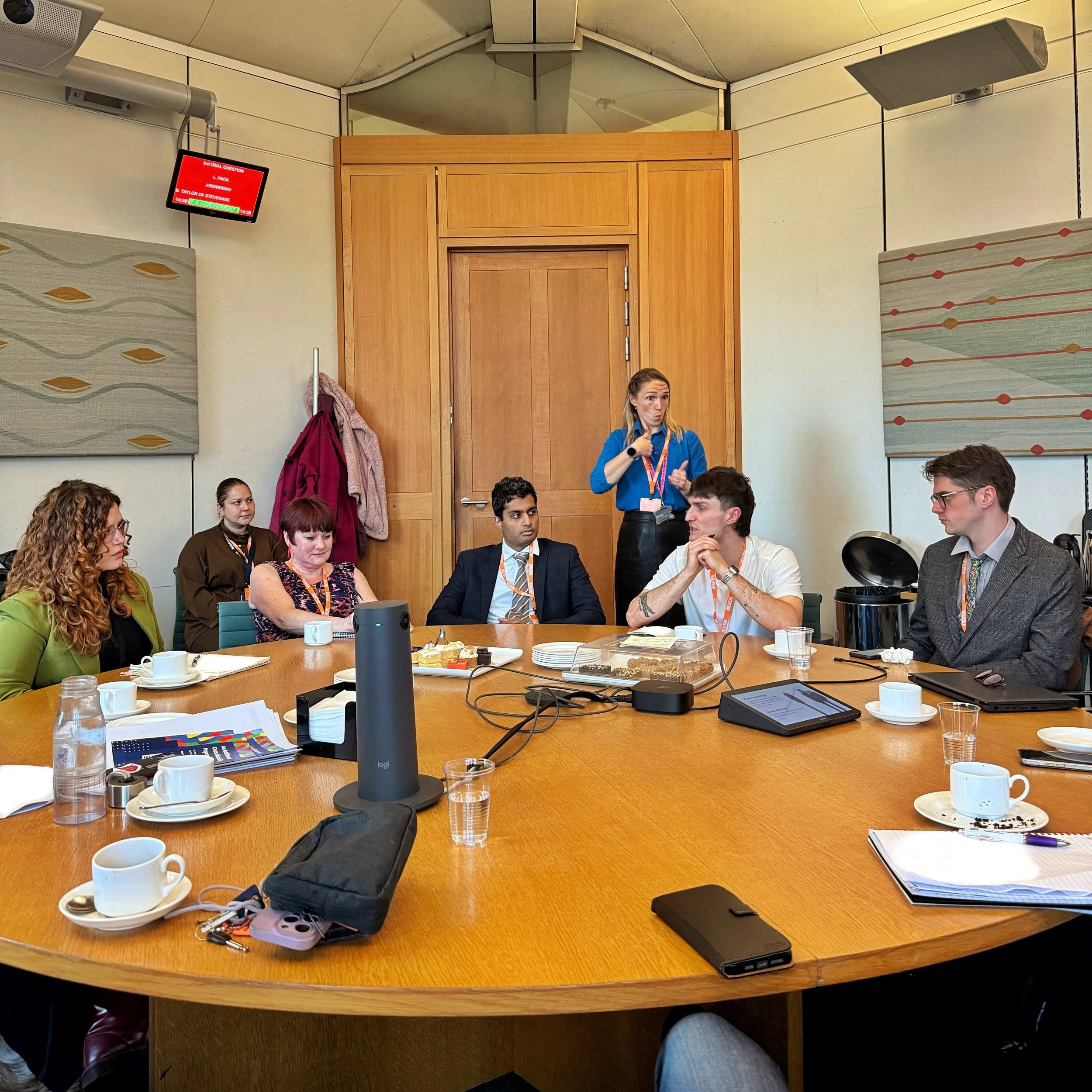 A roundtable discussion in Parliament. From left to right: Bethany, Ellen, Arun and Mikey and Josh engaged in conversation, with a BSL interpreter standing behind. Notes, cakes and laptops are on the table.