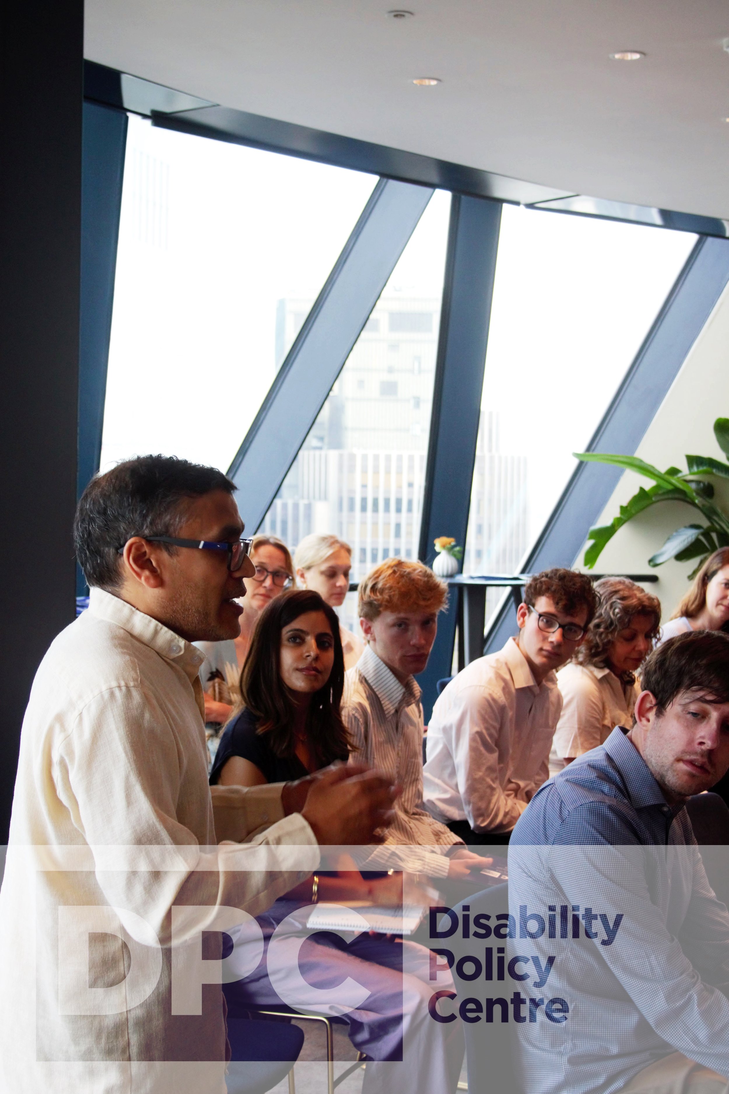 Photograph of Kush stood up at the edge of the audience engaging with the panel. He is wearing a white shirt and glasses. Behind him the audience are sat down with their heads turned to him. At the bottom a DPC logo watermark.