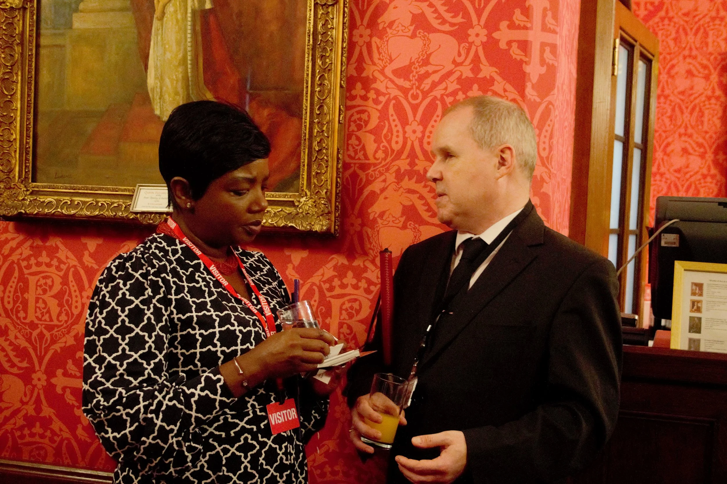 Close up shot of two guests conversating in infront of red wallpaper, on the left is a woman with short black hair in a black and white long sleeved shirt dress with a red visitors langyard on. On the right a man in the black tie suit and white shirt