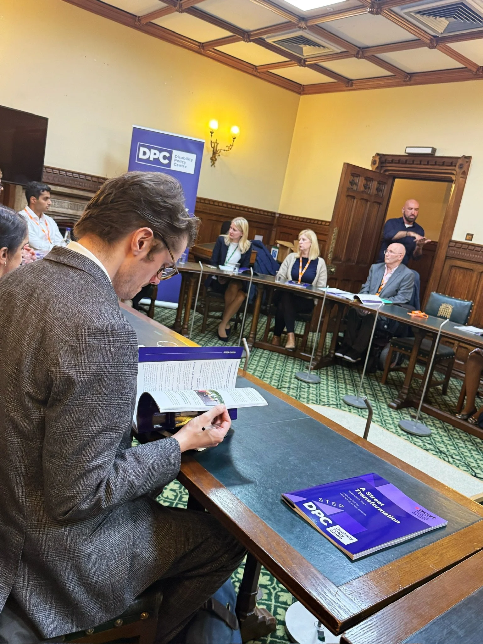 An attendee reading the A Street Transformation report during the panel discussion, with speakers Louise, Ian, and others at the front beside a Disability Policy Centre banner and a sign language interpreter.
