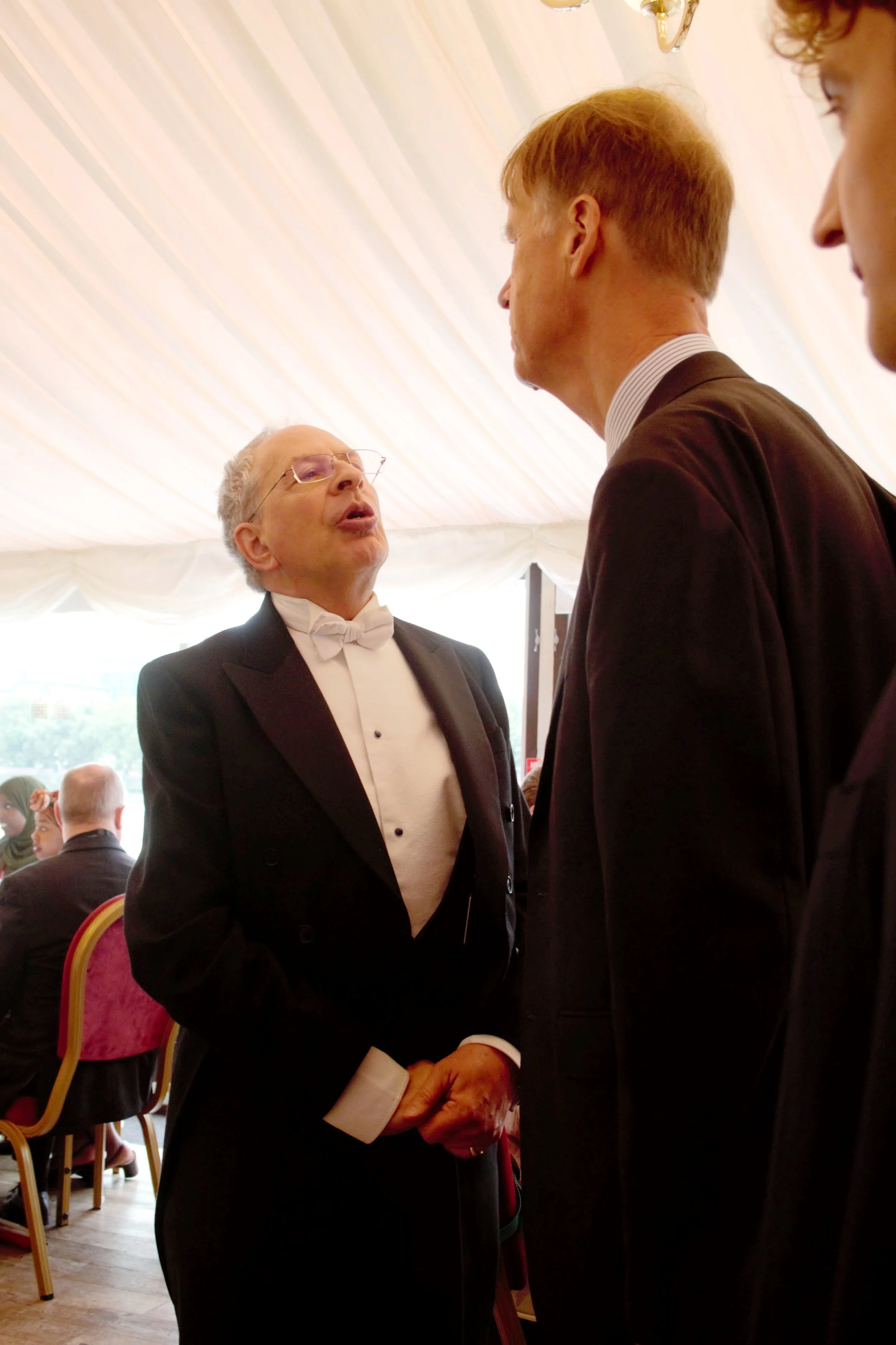 Close up portrait or our host on the left greeting speaker Sir Stephen Timms MP on the right. Host is dressed in a black tail suit with white shirt on bow tie. Sir Timms has his back the camera in a black blazer 