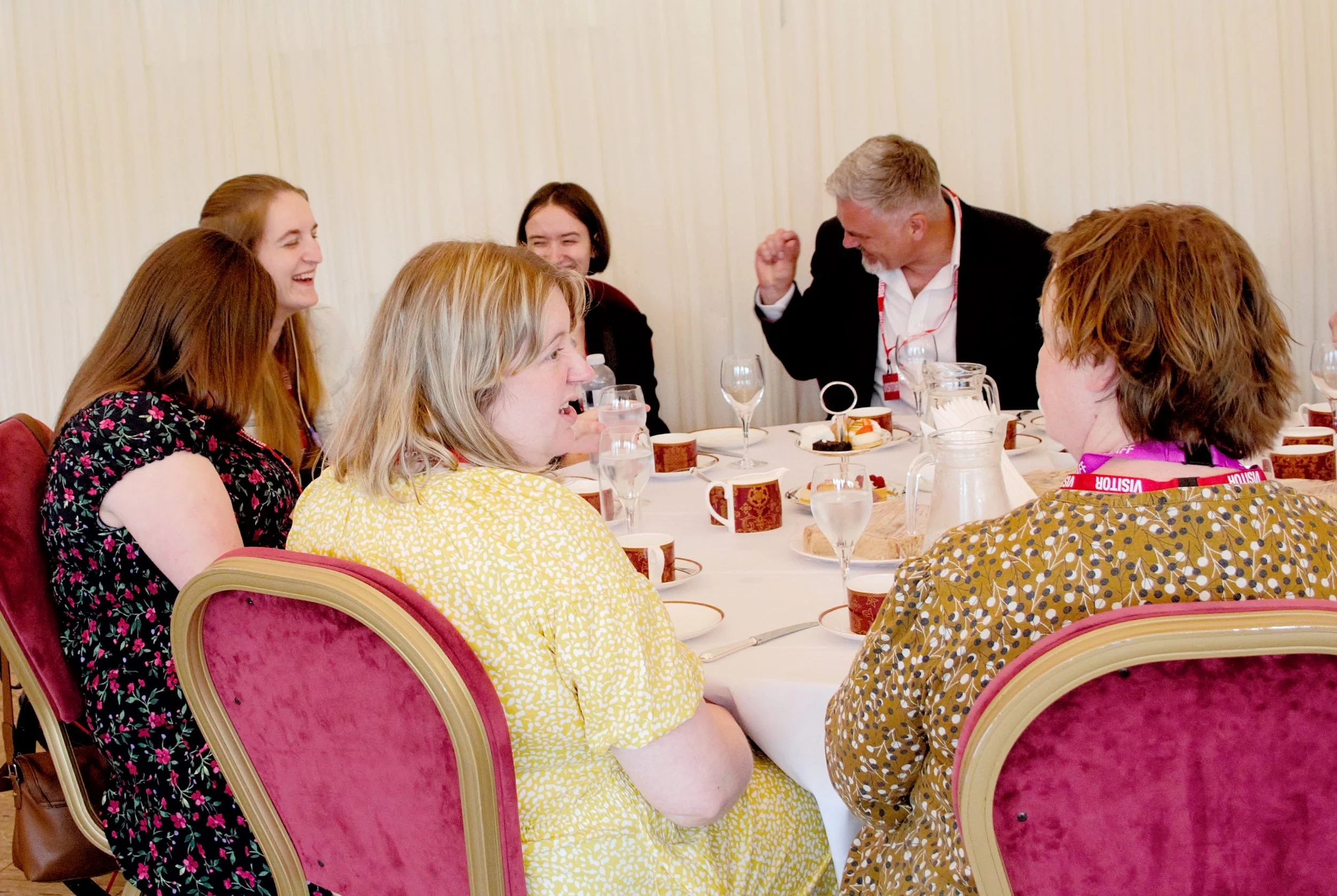 Six guests laughing and socialising around a table of afternoon tea served. Five woman and one man. Three of the guests are woman in floral dresses with their back facing the camera, the man is fist pumping looking to the side dressed in a black suit