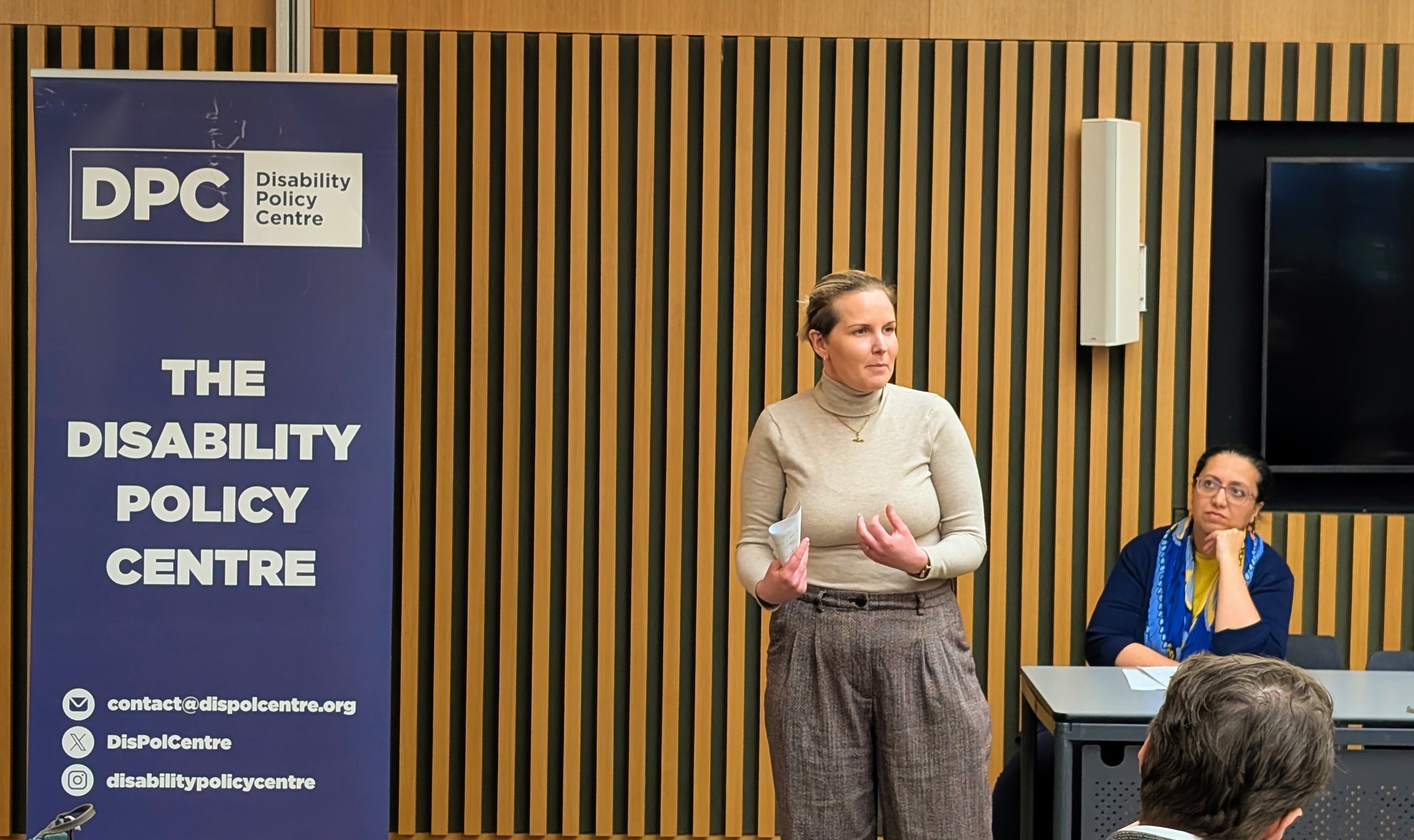 Emma stands speaking at a City Hall roundtable. She has blonde hair and wears a beige jumper, holding notes. Hina, wearing blue, sits beside her listening. A Disability Policy Centre banner stands behind.