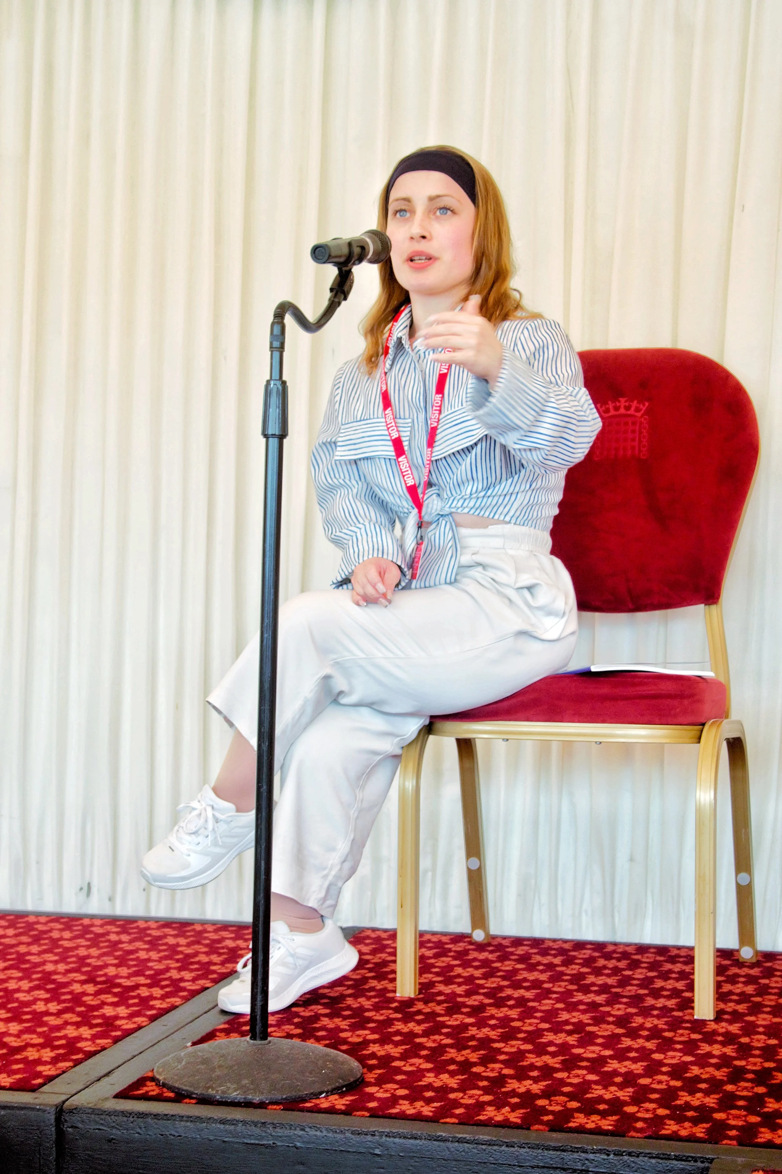Full body of Ellie Robinson MBE, seated on the stage speaking into the microphone. She is dressed in a black and white buttoned up shirt, black headband, white trousers with white trainers and red vistitors langyard 