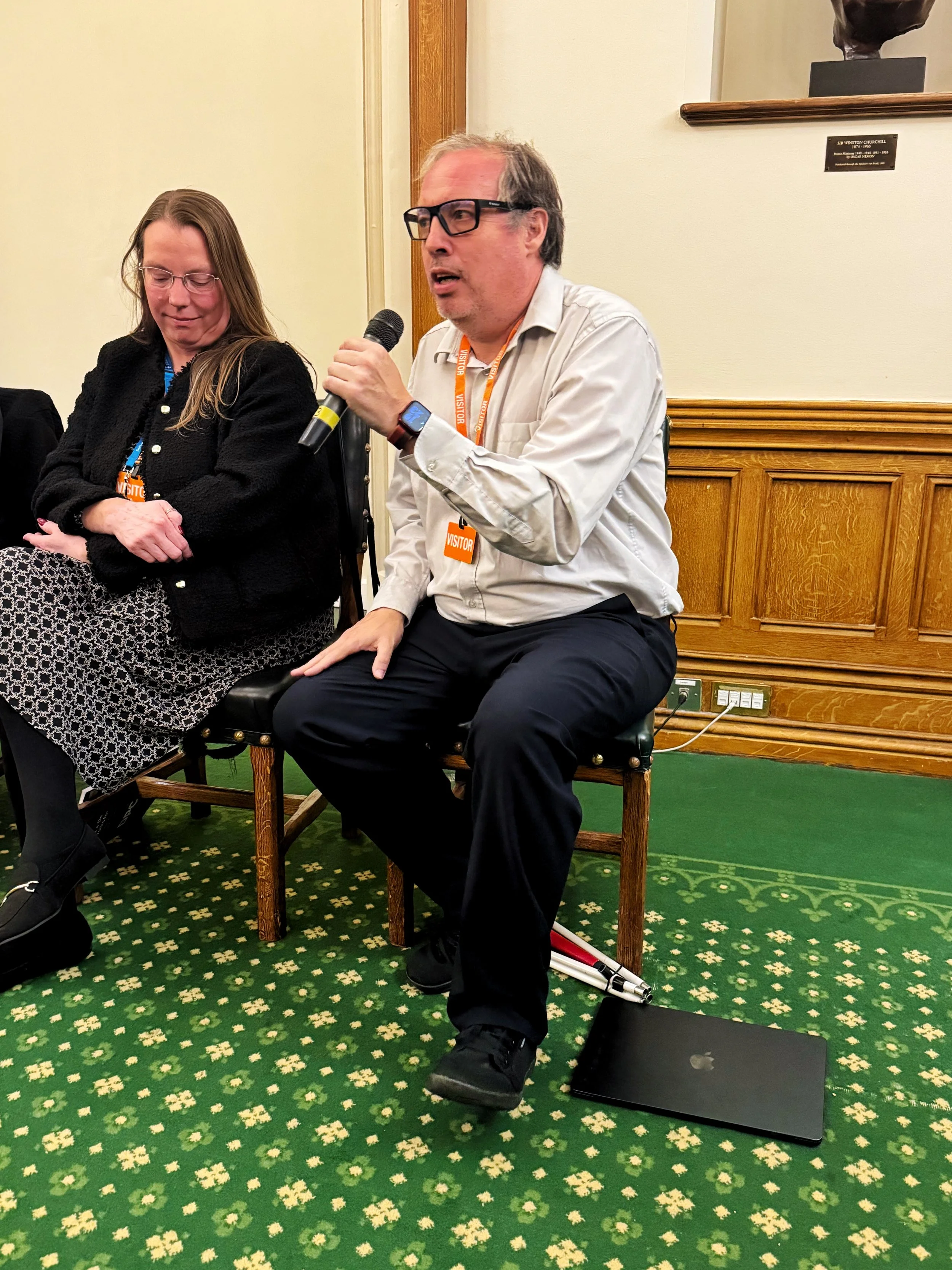 Panel discussion in the Churchill Room, Houses of Parliament, with Mike Wordingham speaking into a microphone beside Michelle Anglesea. A laptop rests on the floor near his feet during the session.