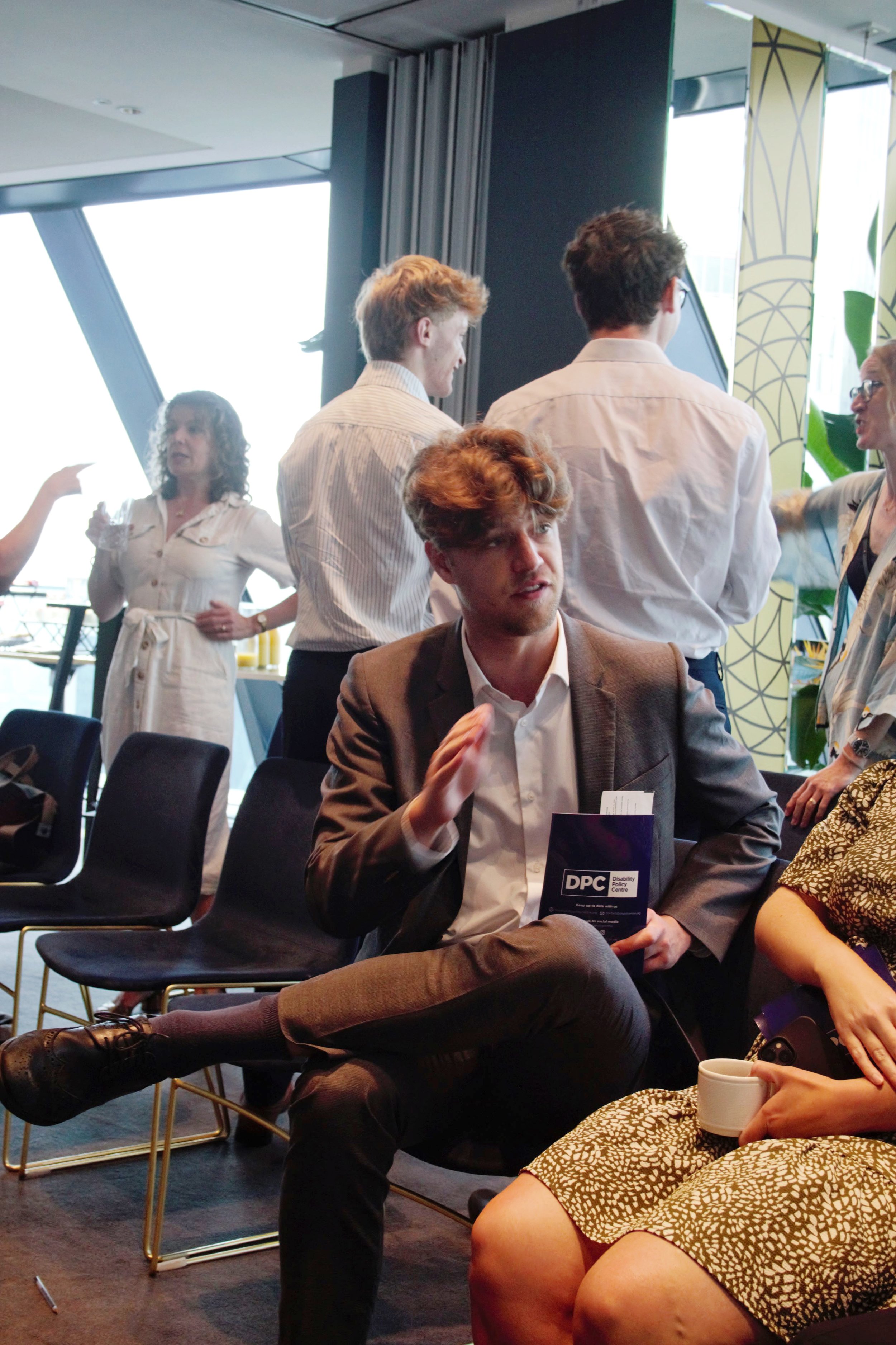Photograph of a male guest sat down holding a printed copy of the DPC report. He is talking and gesturing to a lady sat next to him who's face we can't see. Behind are other attendees stood up and mingling all dressed smartly.