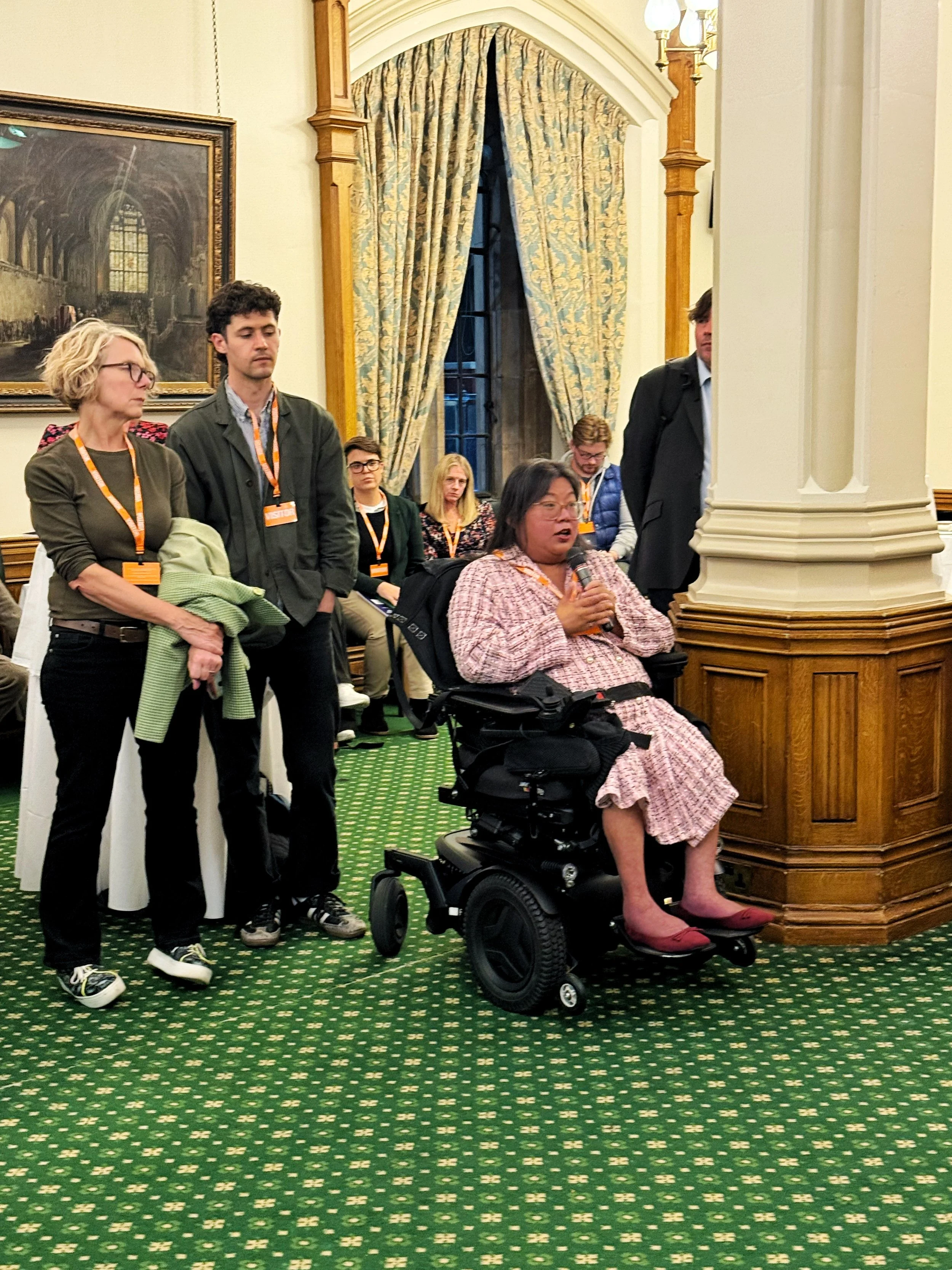 Guest in a wheelchair asks a question during the panel session in the Churchill Room, Houses of Parliament, with other attendees standing and listening in front of tall curtains and paintings.