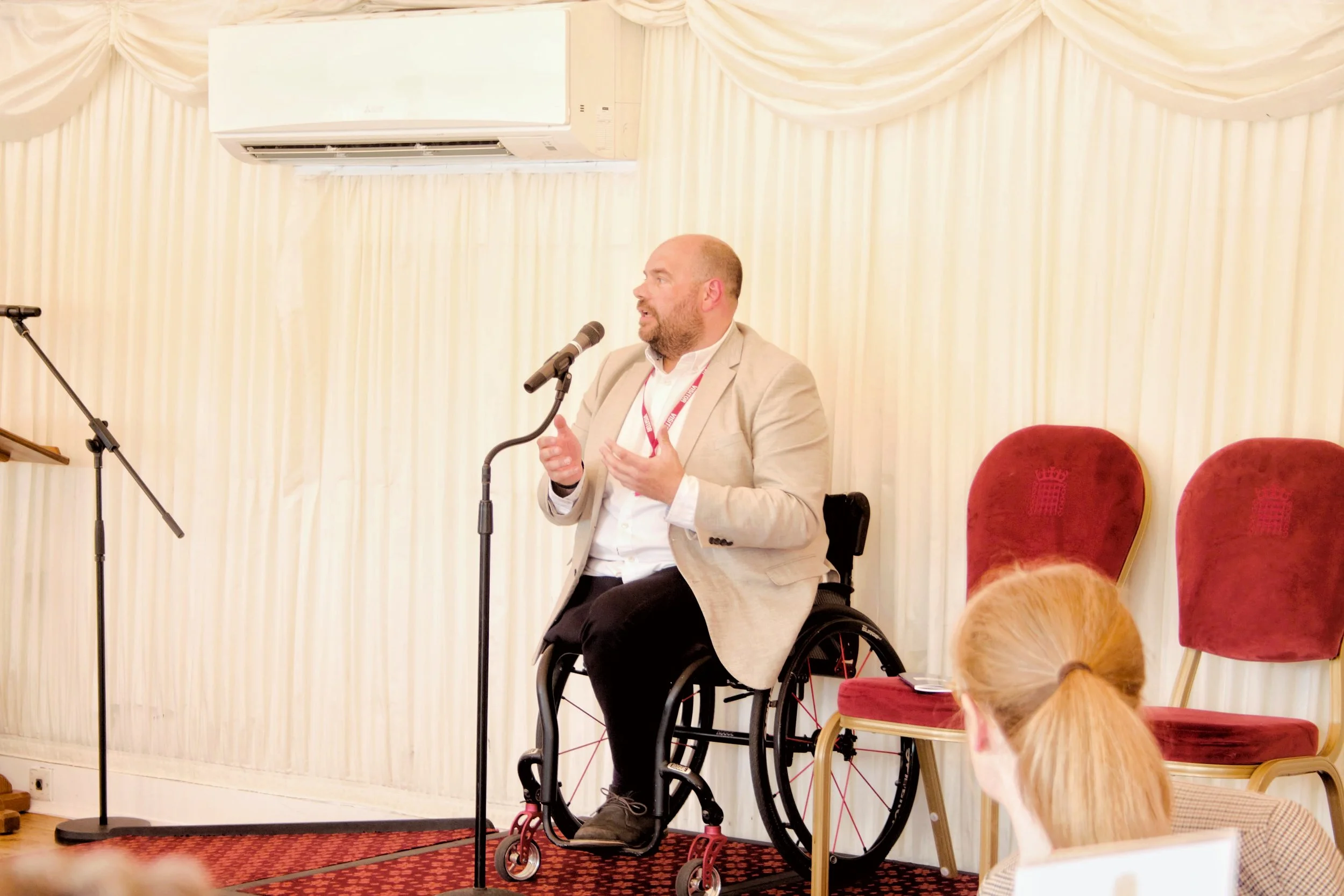 Wide shot from the right of Mark Briggs speaking on the stage into the microphone, he is wearing a white shirt, beige suit jacket, red visiotrs langyard and black trousers , he is in a wheelchair with guests in the forground watching