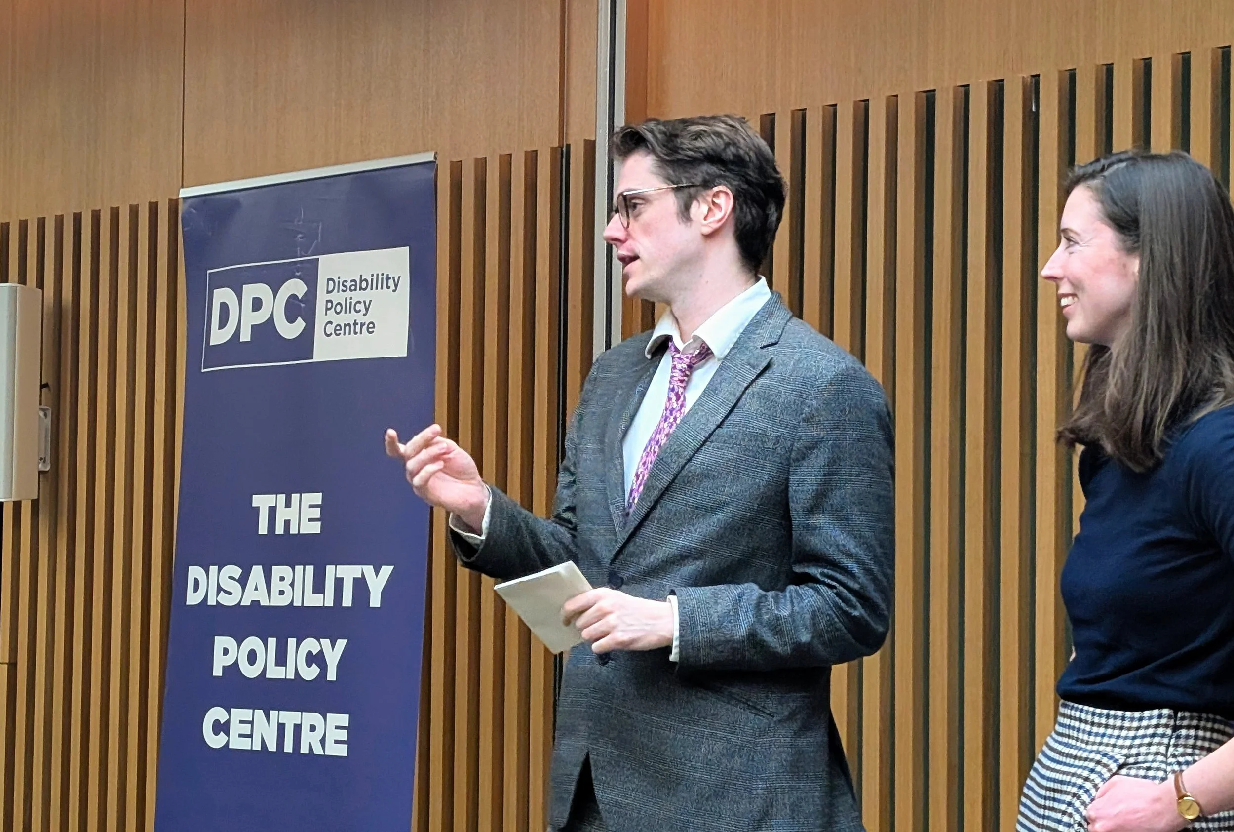 Josh stands speaking at the front of a City Hall room, wearing a grey suit with a red tie, he gesturing to the room and holding papers in one hand. Chloe stands beside him smiling to the right. A Disability Policy Centre banner stand to the left.